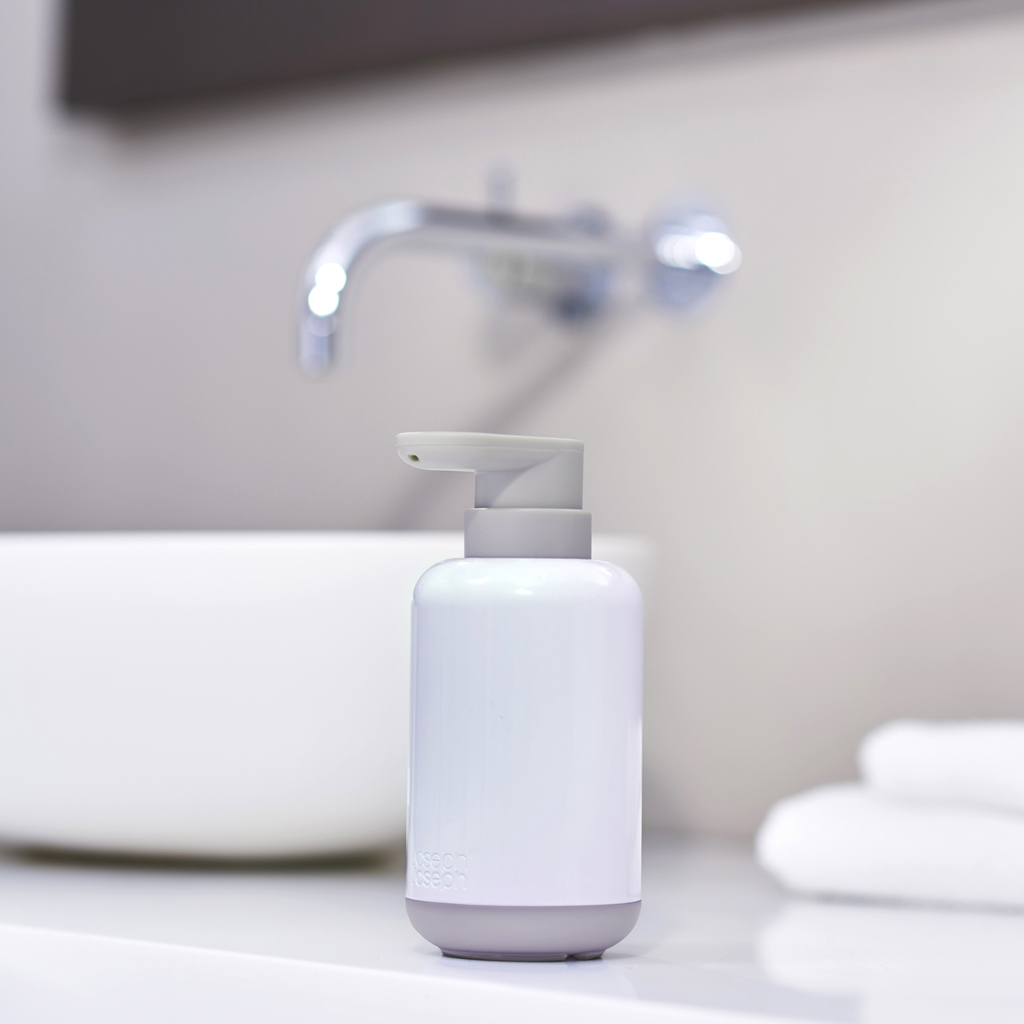 Un dispensador de jabón blanco y gris está sobre una superficie blanca, con un lavabo y grifo borrosos al fondo.