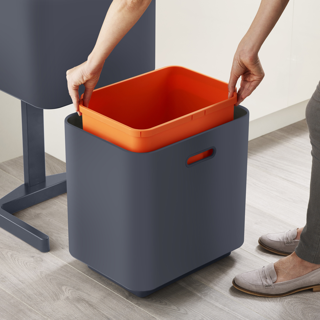 Una persona che mette un contenitore arancione dentro un contenitore blu scuro. Lo sfondo mostra un pavimento in legno e parte di un muro bianco.