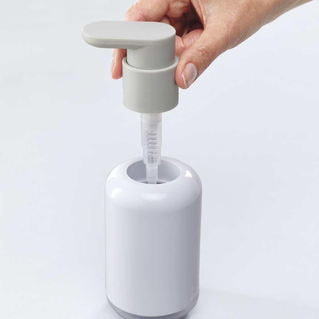 Una mano presiona una bomba gris en un dispensador de jabón blanco. El dispensador está sobre una superficie blanca.
