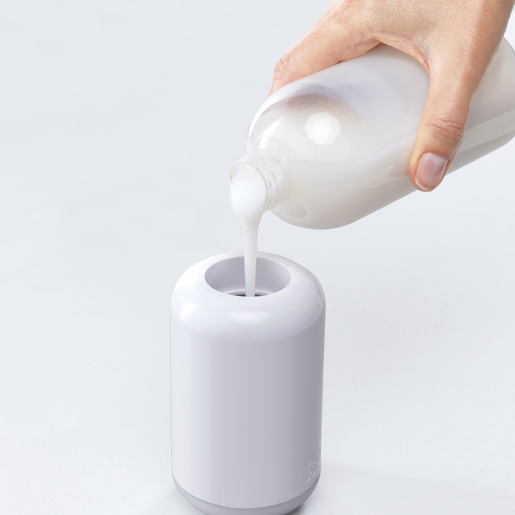 Una mano vierte líquido blanco de una botella en un dispensador blanco. El dispensador está sobre una superficie blanca. El fondo es blanco.