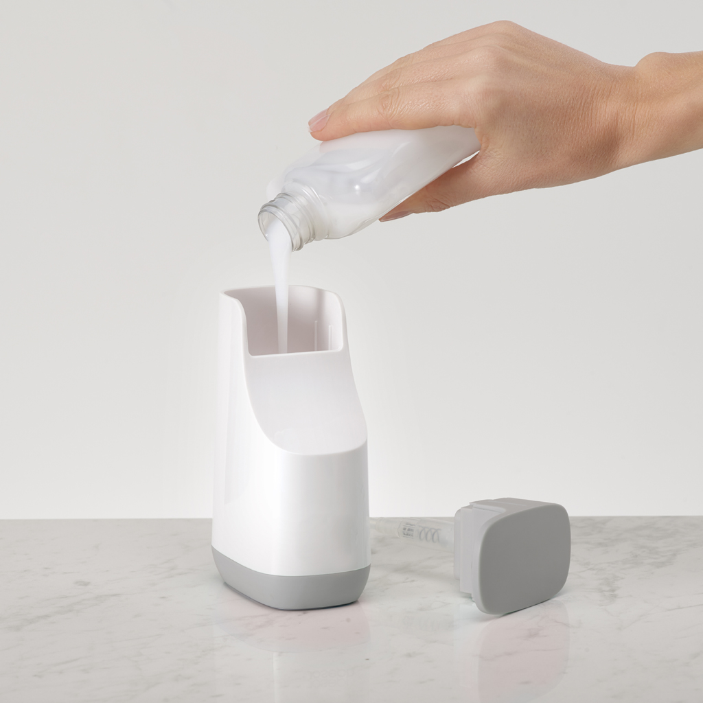 Una mano vierte líquido blanco de una botella en un dispensador blanco con base gris, junto a un componente cuadrado gris.