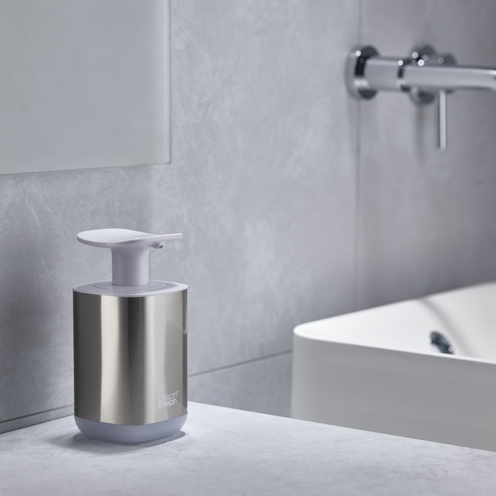 Un dispensador de jabón metálico con tapa blanca sobre encimera gris. Un lavabo blanco y grifo en el fondo, contra una pared gris.