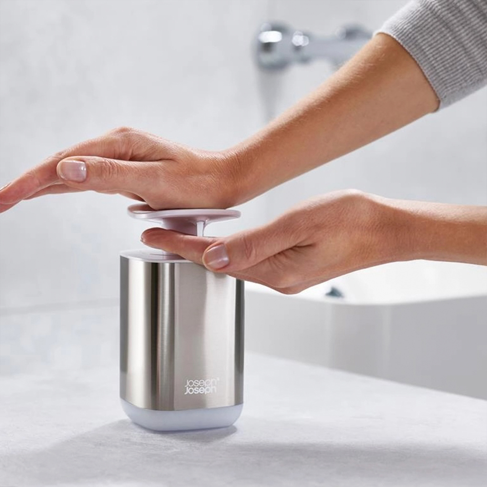 Una persona presionando un dispensador de jabón con las manos. El dispensador es plateado con una tapa blanca. Un lavabo está en el fondo.