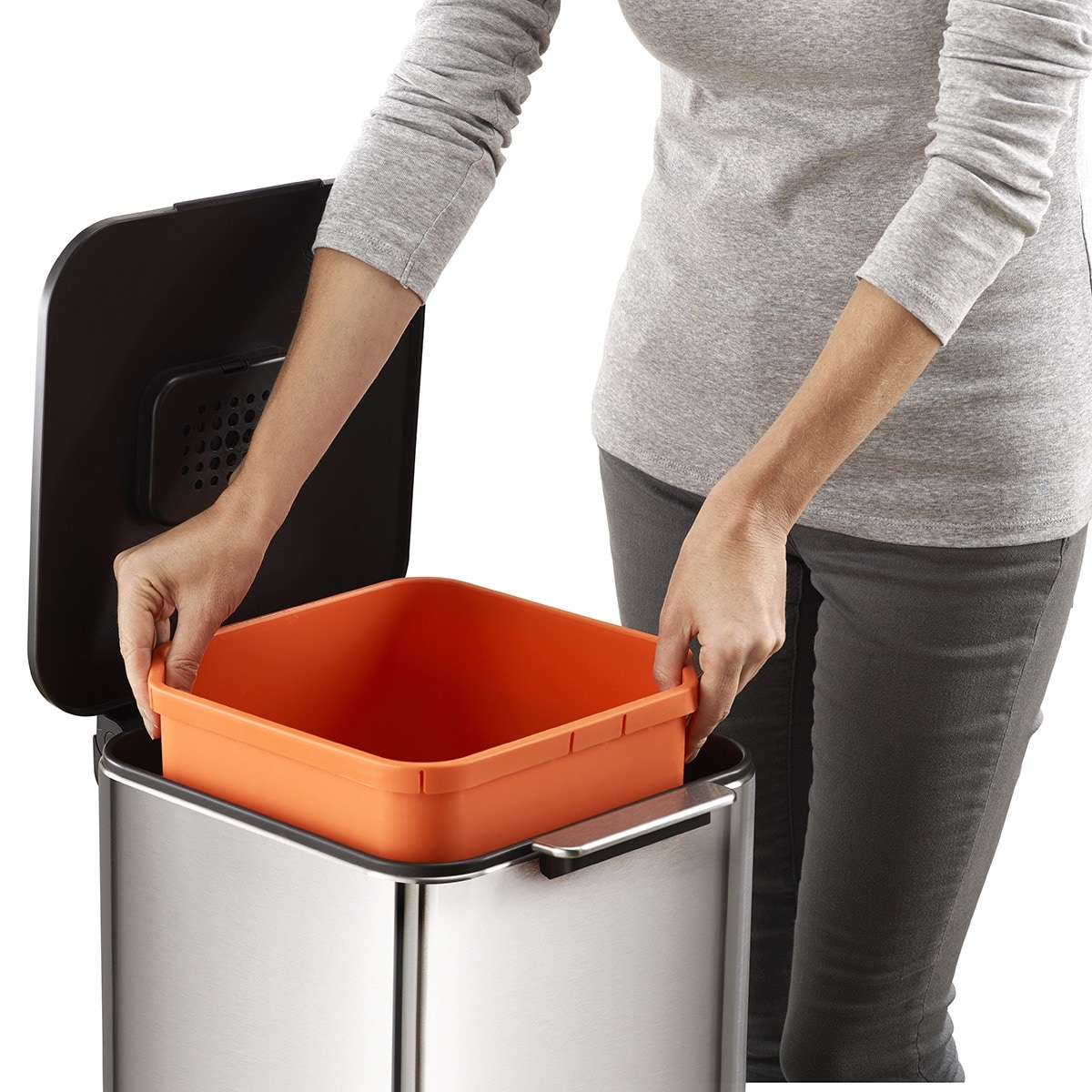 Una persona sostiene un cubo de basura naranja dentro de un cubo de basura de acero inoxidable. La tapa es negra y está abierta.