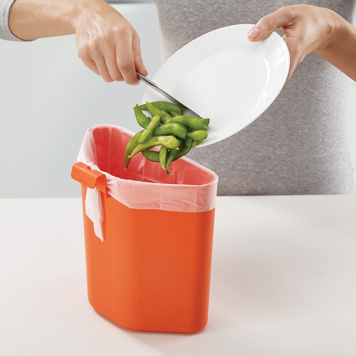 Una persona tira edamame de un plato blanco a un cubo de basura naranja. El cubo de basura tiene un forro blanco.