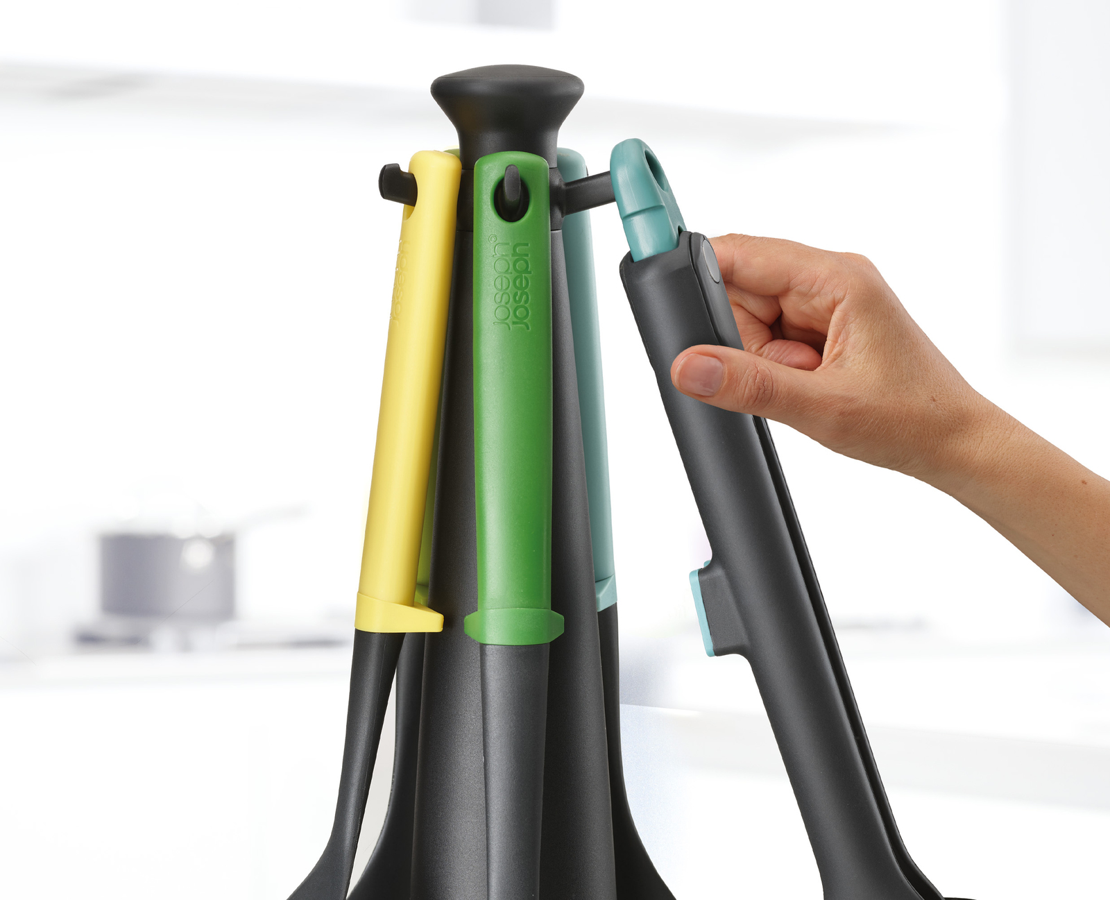 Una mano sostiene un utensilio de cocina negro de un conjunto de herramientas coloridas sobre un soporte negro. Se ven herramientas amarillas, verdes y turquesas.