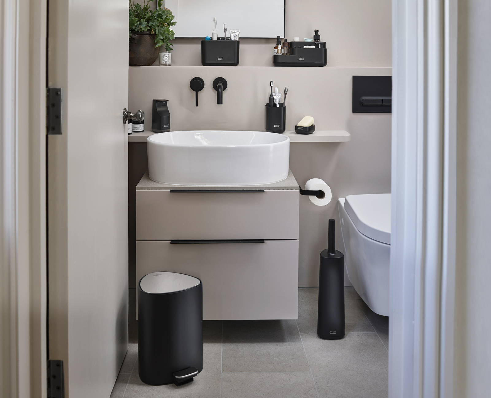 Un baño moderno con lavabo blanco, accesorios negros y un bote de basura negro. Artículos de aseo en un estante.