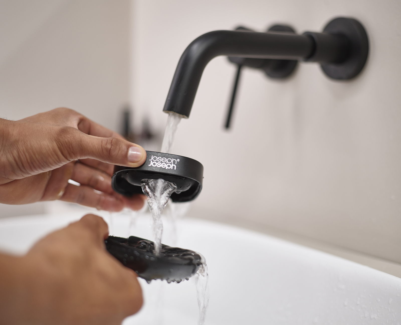 Manos sosteniendo un producto negro Joseph Joseph debajo de un grifo abierto. El agua fluye hacia el producto y debajo hay un lavabo blanco.