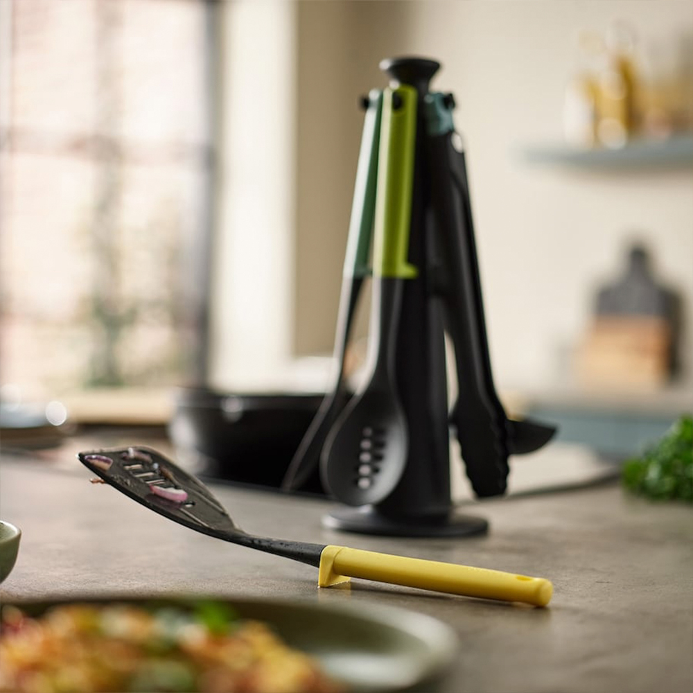 Utensilios de cocina en una encimera. Una espátula negra con mango amarillo en primer plano. Un soporte para utensilios en el fondo.