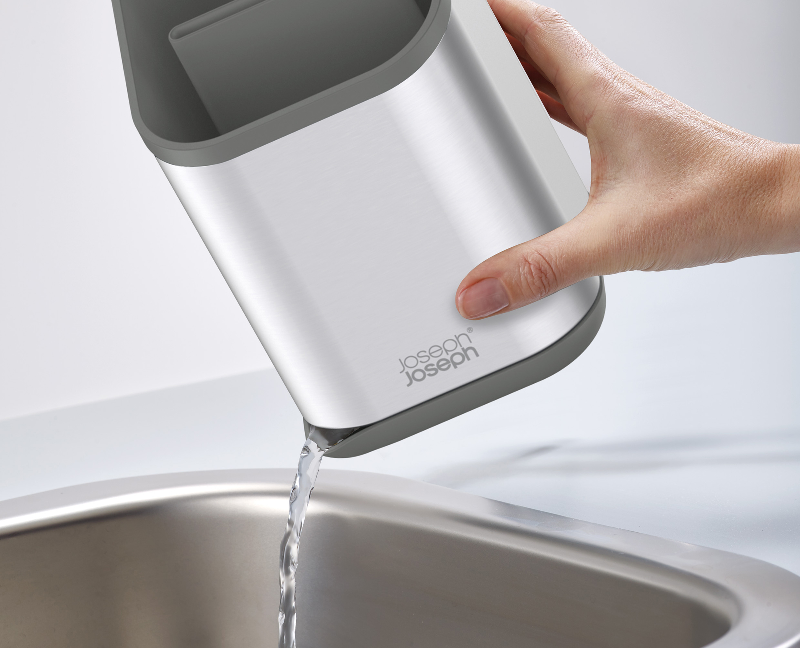 Una mano versa acqua da un contenitore argento e grigio in un lavandino in acciaio inox. Il contenitore ha un logo Joseph Joseph.