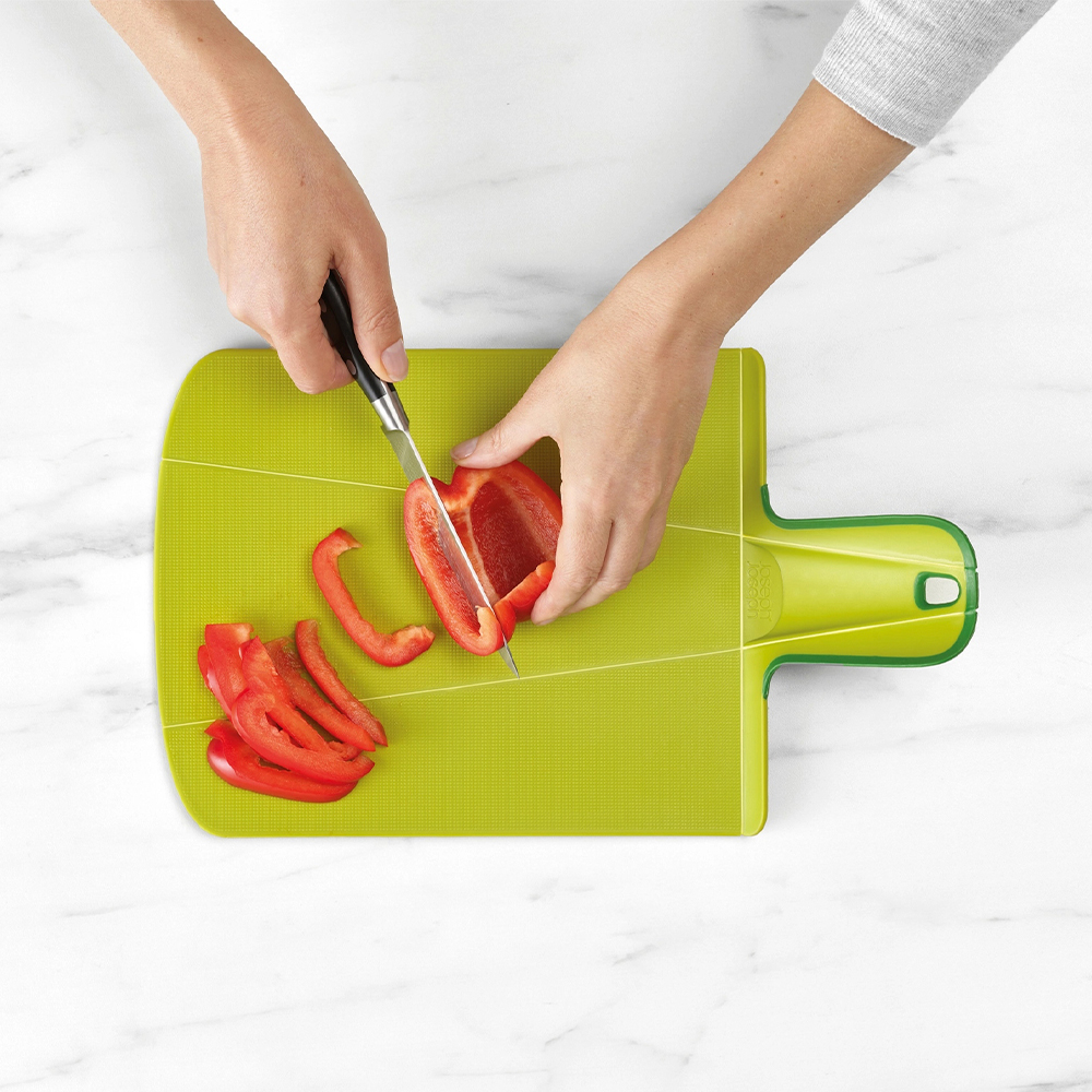 Manos cortando pimiento rojo sobre tabla verde. Trozos cortados a la izquierda. Fondo de mármol blanco.