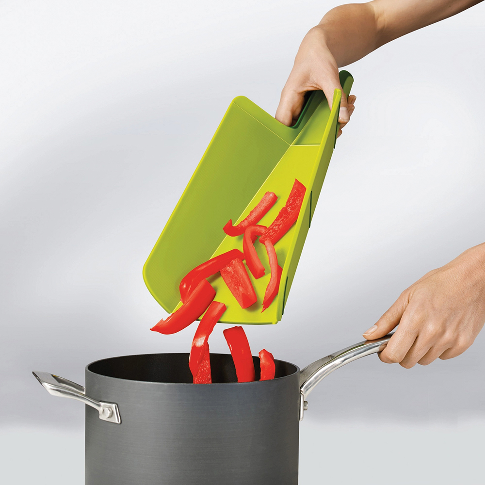 Manos vertiendo rodajas de pimiento rojo de una tabla de cortar verde en una olla. La olla está sobre una superficie blanca.