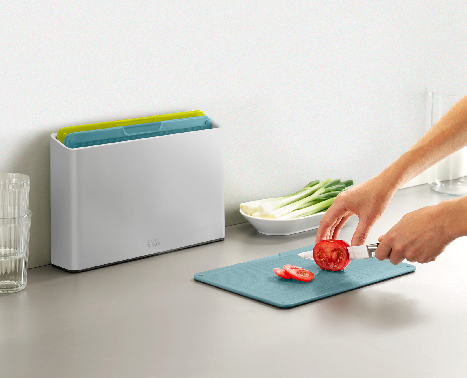 Una persona corta un tomate en una tabla de cortar azul. Un recipiente blanco contiene tablas de cortar. Cebolletas en un bol.