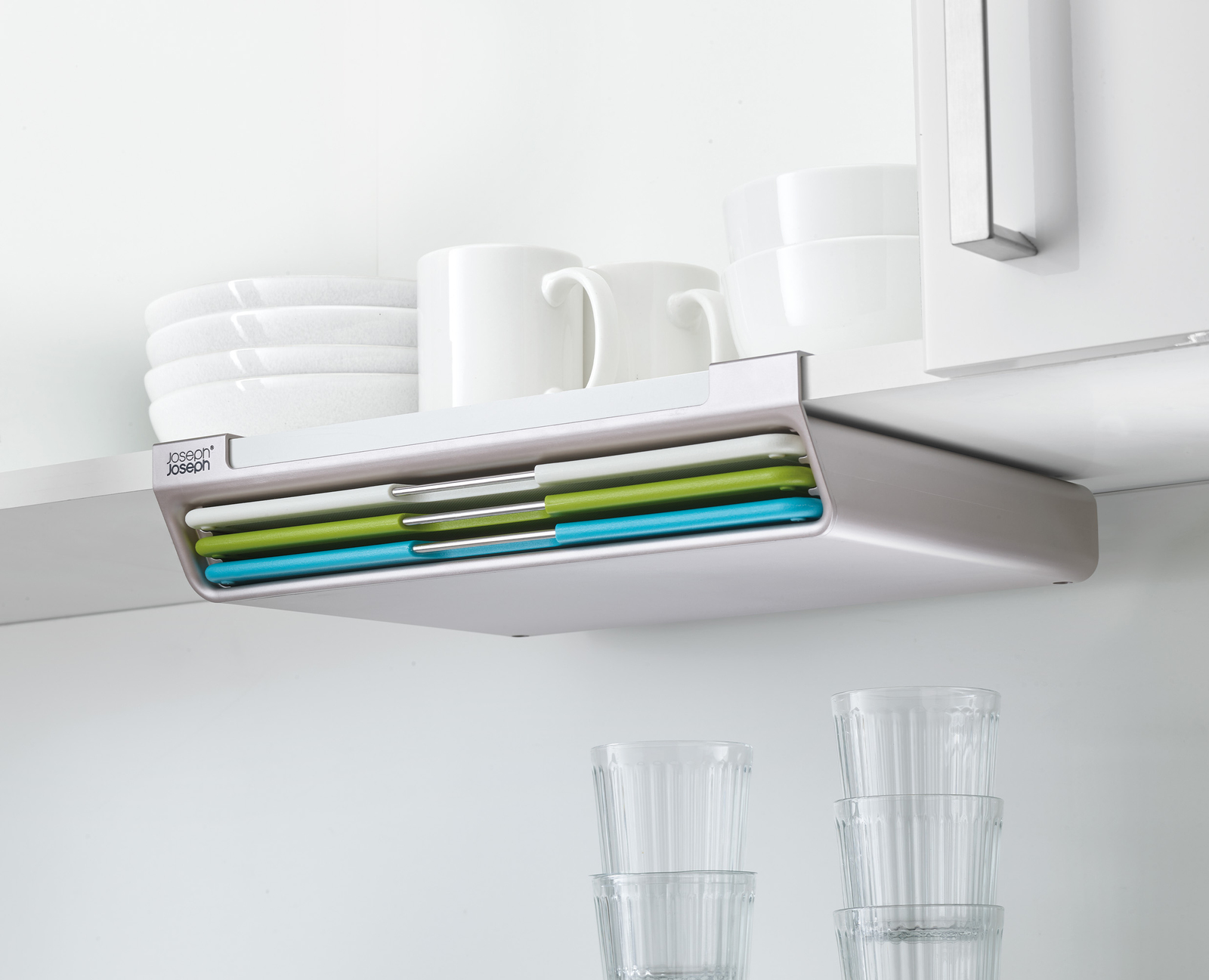Estante de cocina con platos, tazas y tablas de cortar blancos. Tablas verdes, azules y blancas en un soporte. Vasos transparentes debajo.