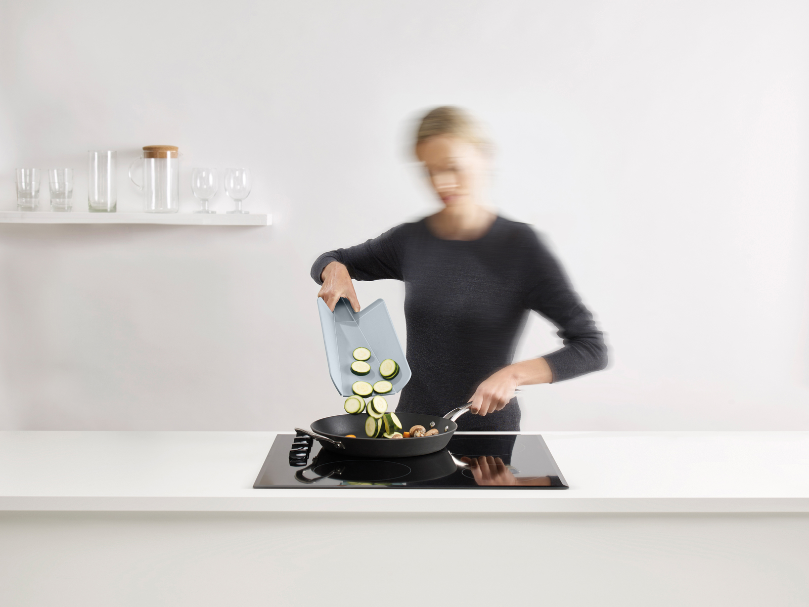 Una mujer agrega calabacín en rodajas a una sartén en una estufa. La cocina tiene un diseño minimalista.