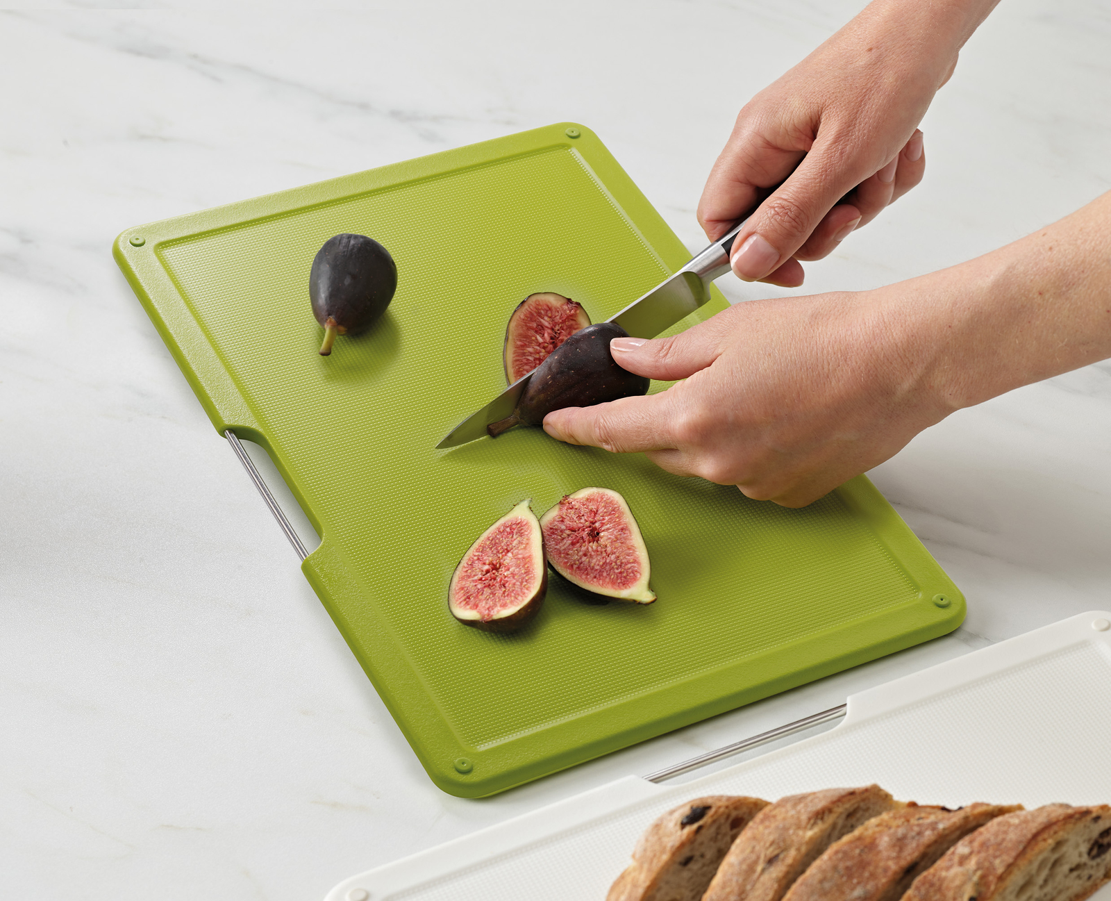 Manos cortan un higo sobre una tabla de cortar verde. También hay higos cortados y enteros. El pan es visible en el fondo.