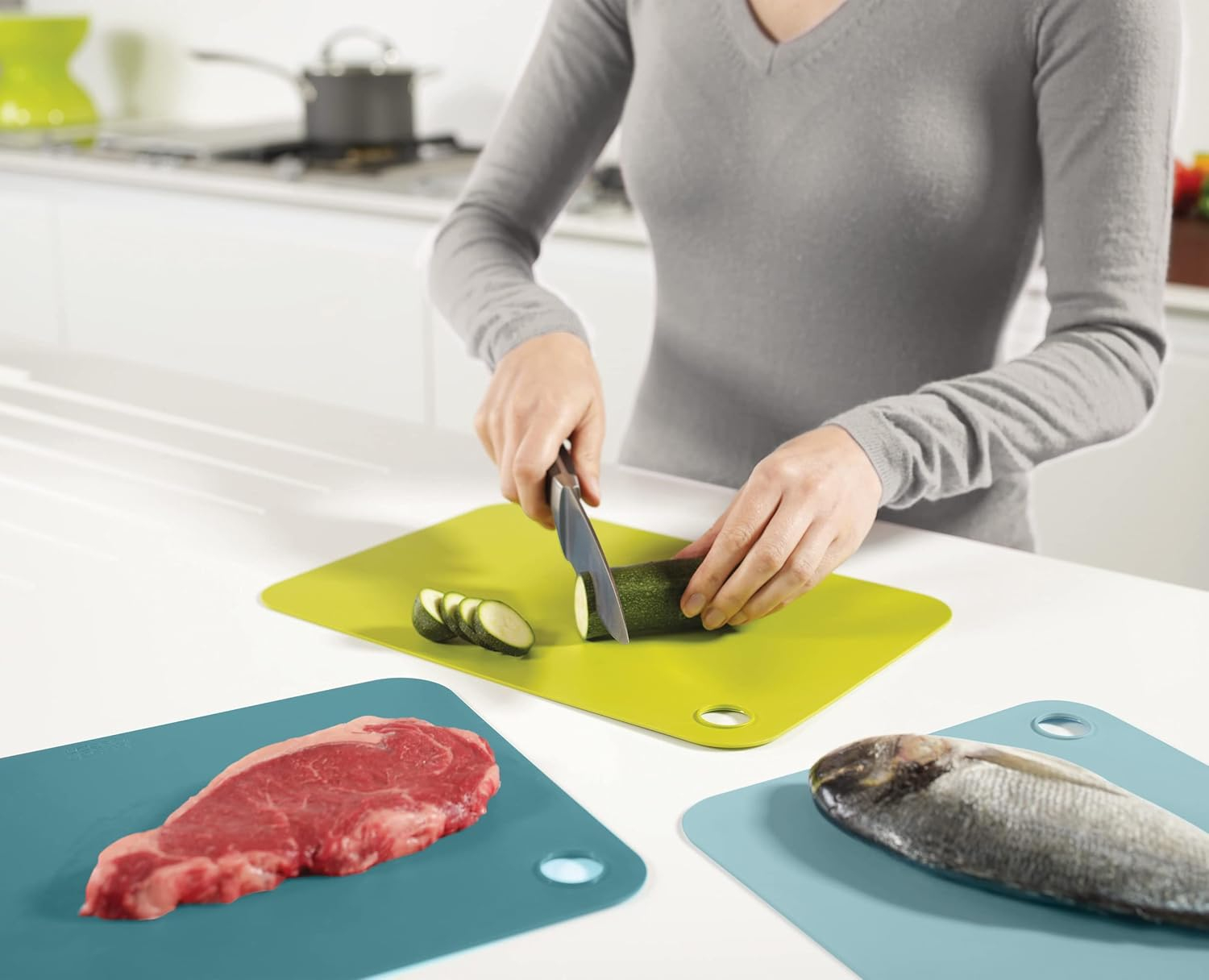 Una persona corta calabacín en una tabla verde. Carne cruda y pescado descansan en tablas azules en una cocina luminosa.