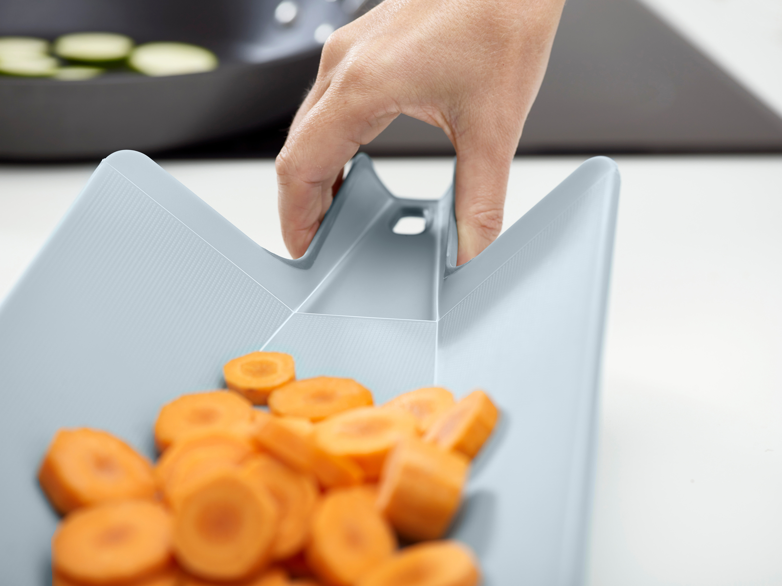 Una mano sostiene una tabla de cortar gris con rodajas de zanahoria naranja. Rodajas de calabacín en una sartén al fondo.
