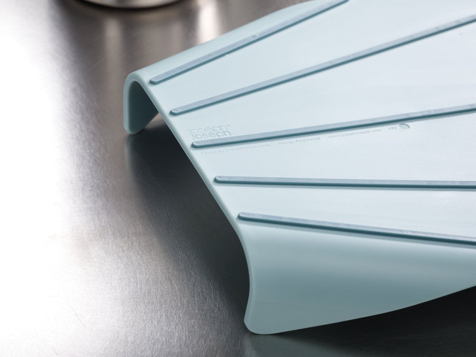 Un utensilio de cocina curvo de color azul claro sobre una superficie metálica. Tiene crestas y el logotipo de Joseph Joseph.