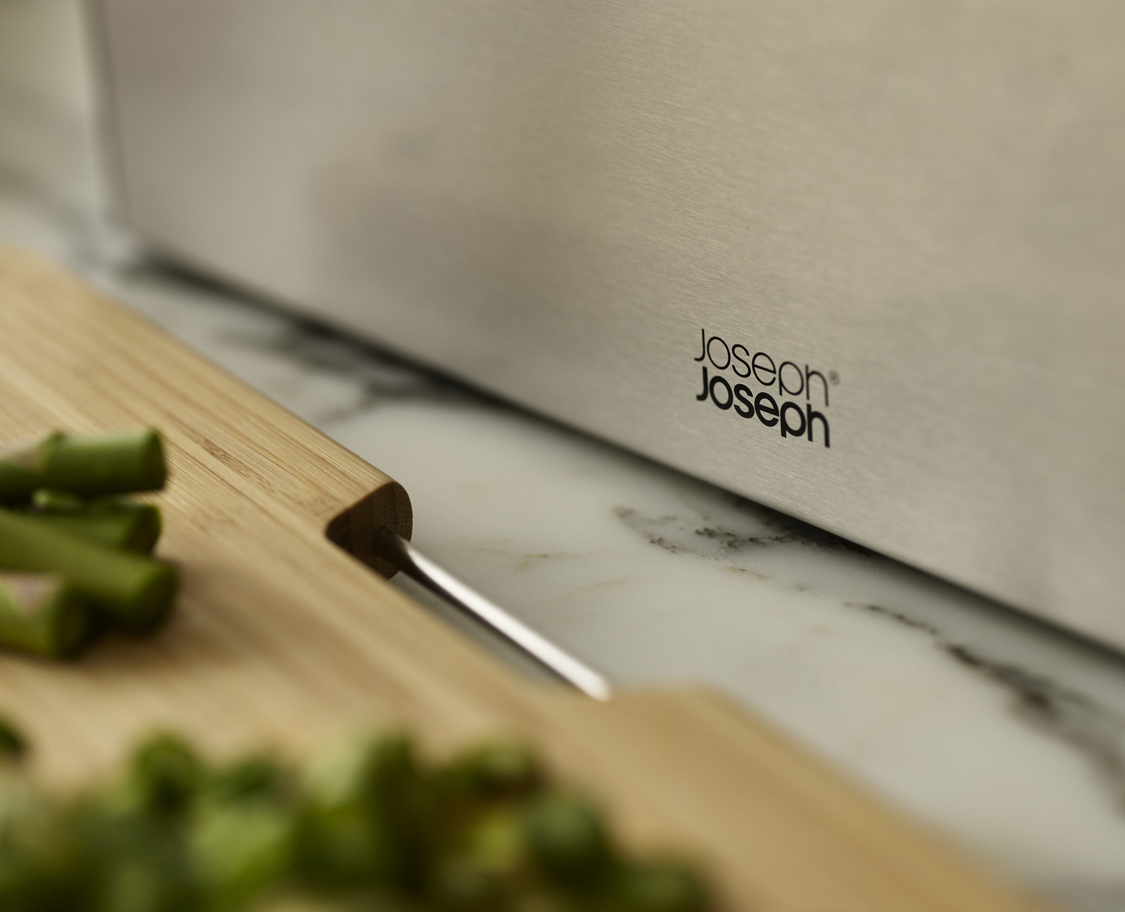 Primer plano de un producto Joseph Joseph. Una tabla de cortar de madera con verduras verdes y una superficie de acero inoxidable con el logo.