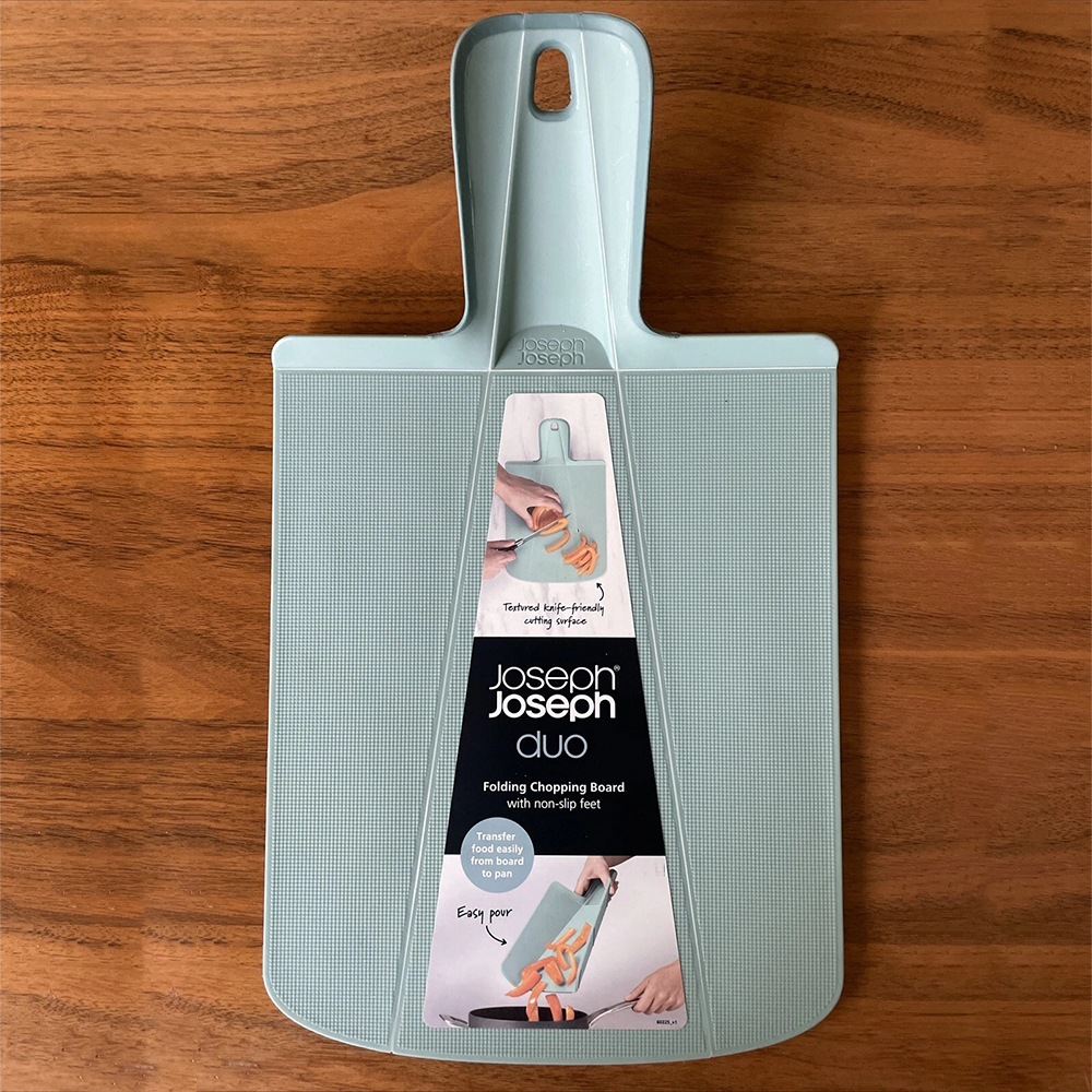Una tabla de cortar plegable Joseph Joseph Duo azul claro sobre una superficie de madera. La tabla tiene un asa y detalles del producto.