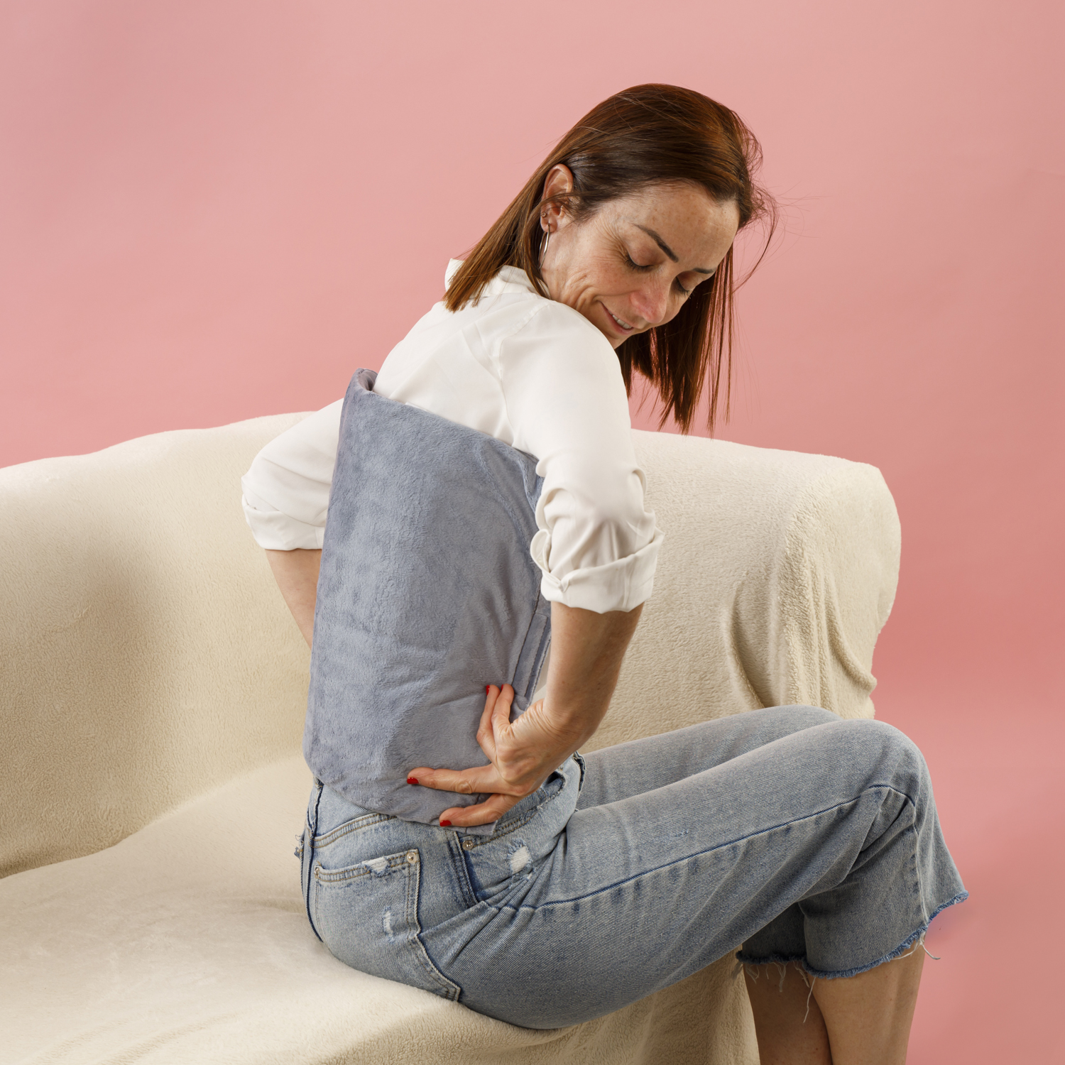 Mujer de pelo castaño sentada en un sofá beige, sosteniendo una almohada azul contra su espalda. Fondo rosa.