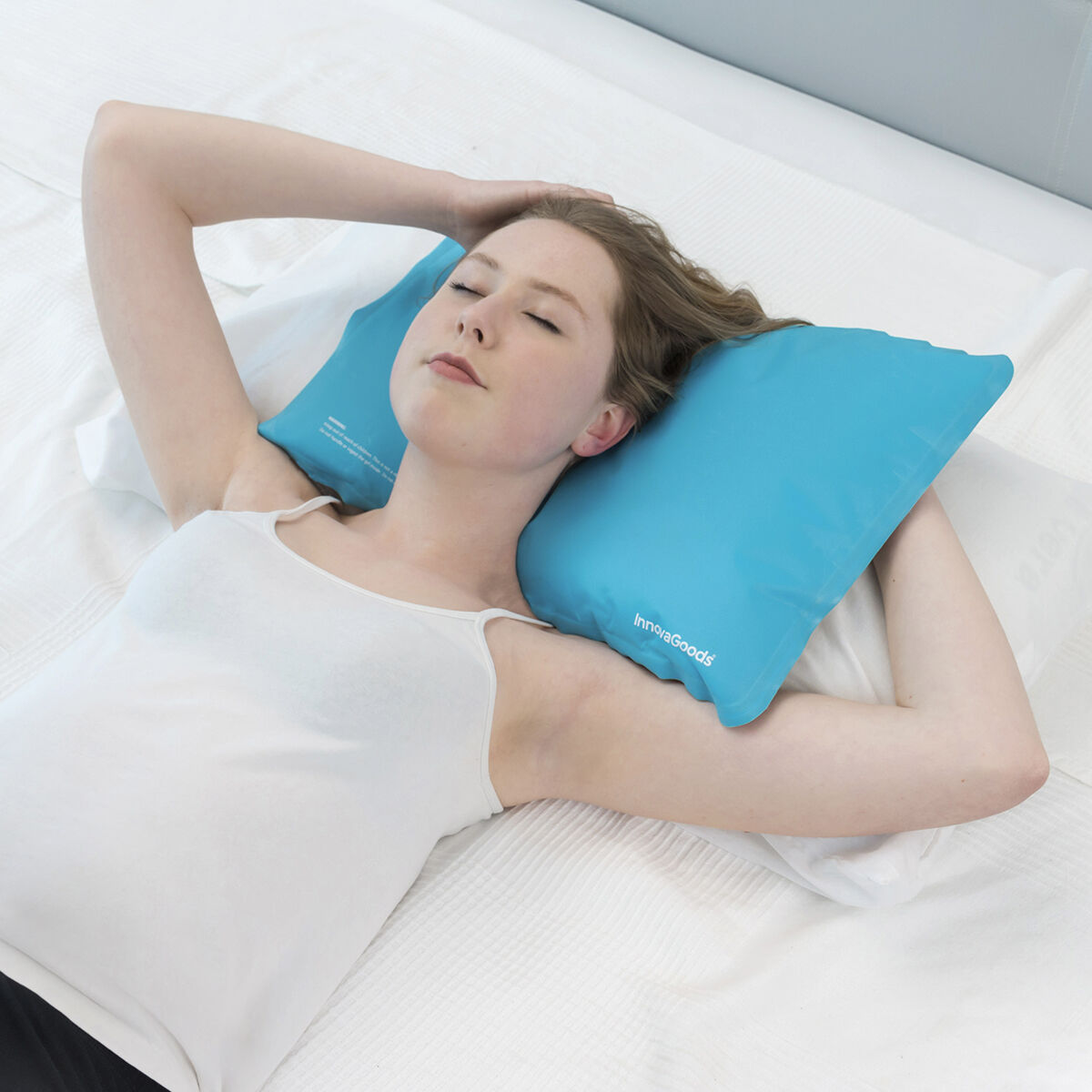 Una donna è sdraiata su un letto con un cuscino blu. Indossa una canotta bianca, mani dietro la testa, occhi chiusi.