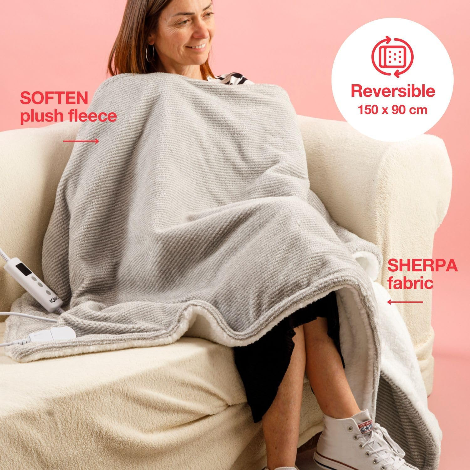 Una mujer sentada en un sofá beige envuelta en una manta gris. La manta es reversible con tejido sherpa. Viste top negro y zapatos blancos.