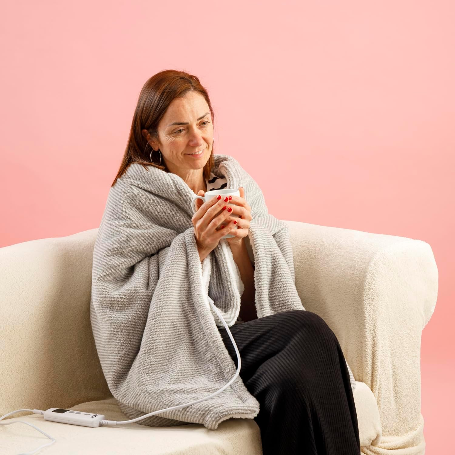 Una mujer sentada en un sofá, envuelta en una manta gris, sosteniendo una taza blanca. Sonríe, con un fondo rosa.