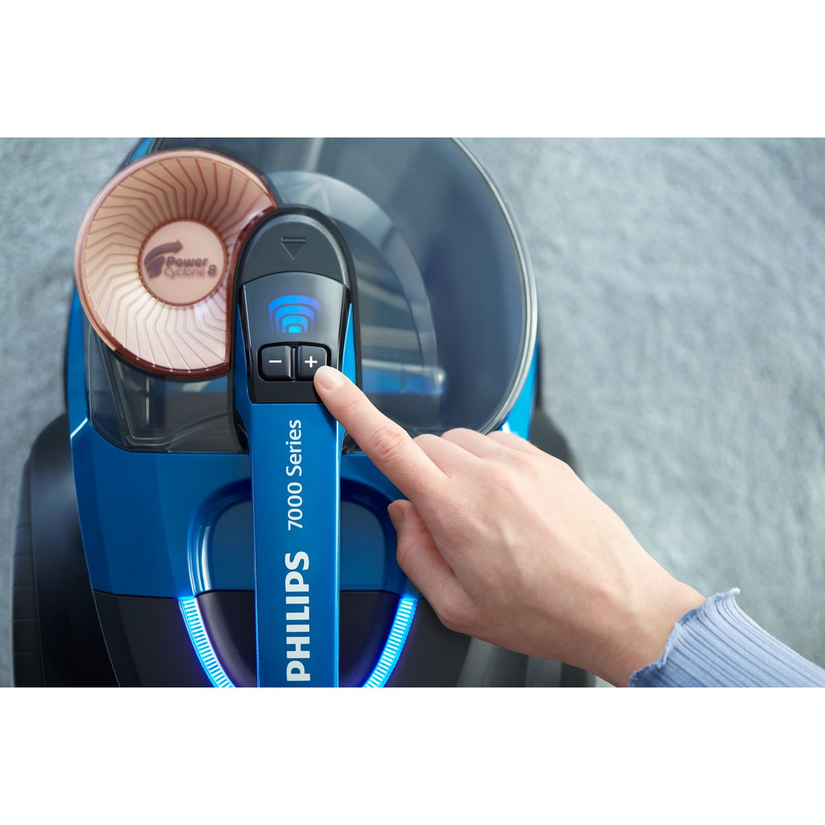 Eine Hand drückt die Plus-Taste auf einem blauen Philips-Staubsauger. Der Staubsauger hat einen Zyklonfilter und befindet sich auf einer grauen Oberfläche.