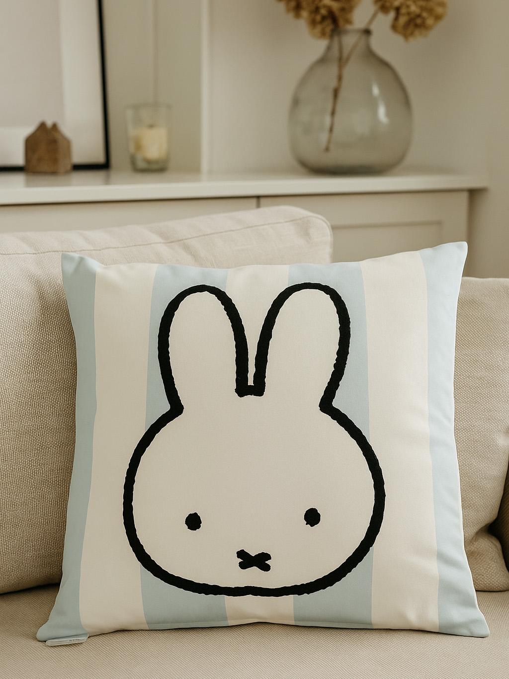 Ein Kissen mit Hasengesicht liegt auf einem Sofa. Das Kissen hat hellblaue Streifen. Eine Vase und eine Kerze stehen auf dem Regal.