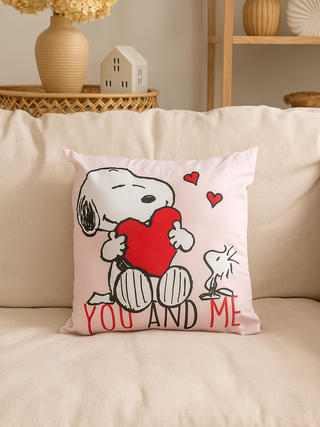 Ein rosa Kissen zeigt Snoopy, der ein rotes Herz umarmt, mit dem Text 'YOU AND ME'. Zwei Herzen schweben darüber. Das Kissen liegt auf einem beigen Sofa.