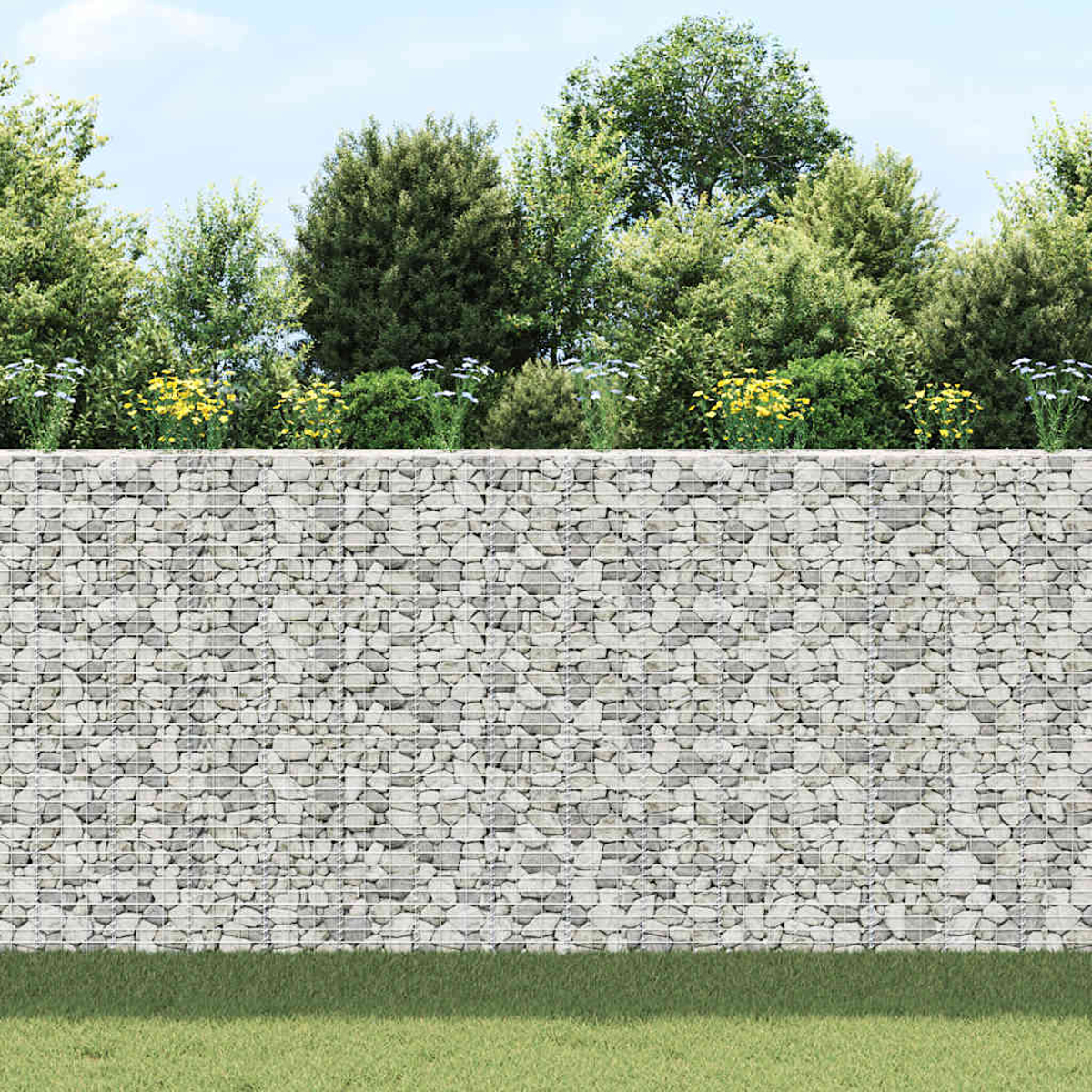 Eine Steinmauer mit Drahtgeflecht, Gras davor und grüne Bäume mit gelben und weißen Blumen oben, unter blauem Himmel.