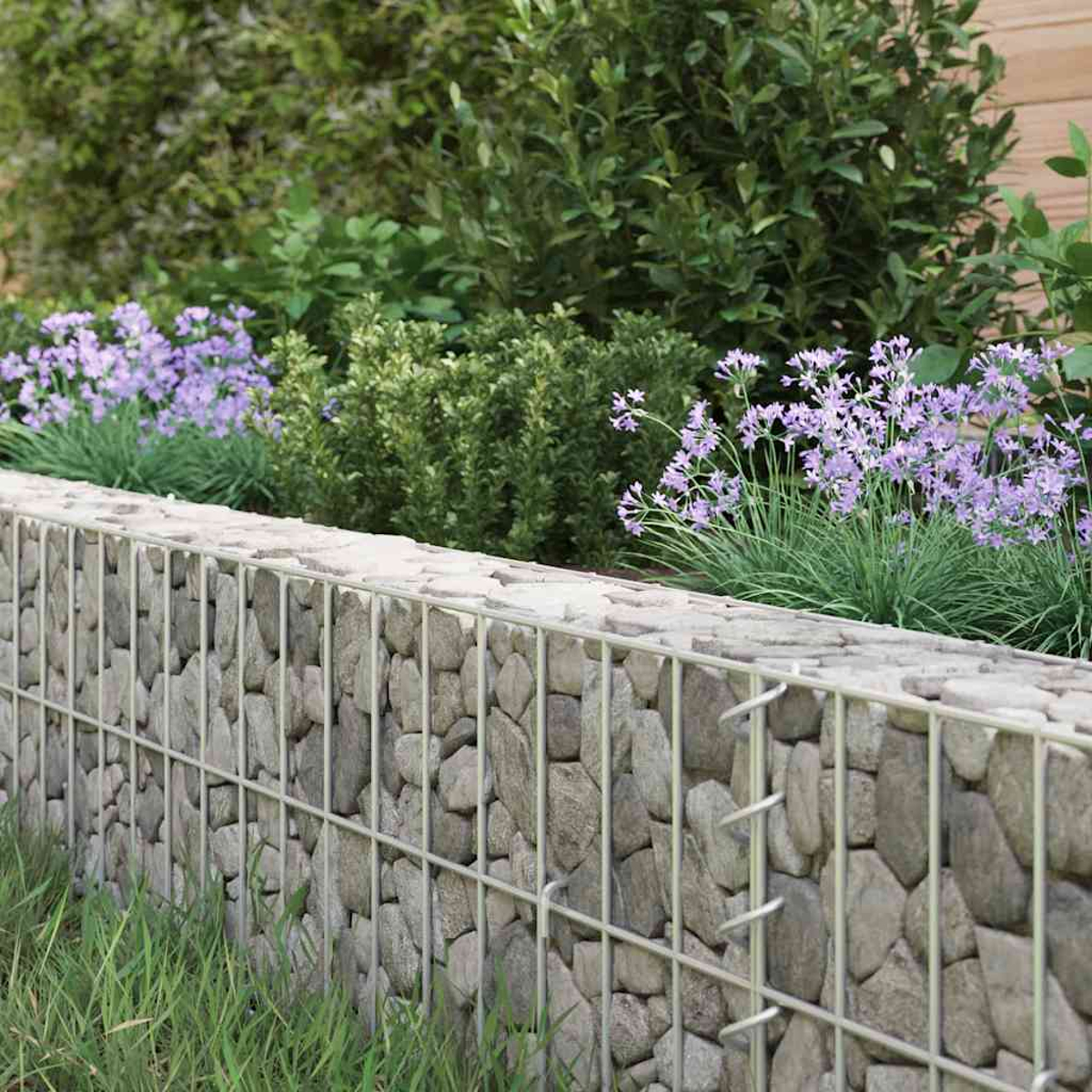 Eine Steinmauer mit Metallgitter, lila Blumen, grüne Büsche und Gras davor. Der Hintergrund hat mehr Grün.