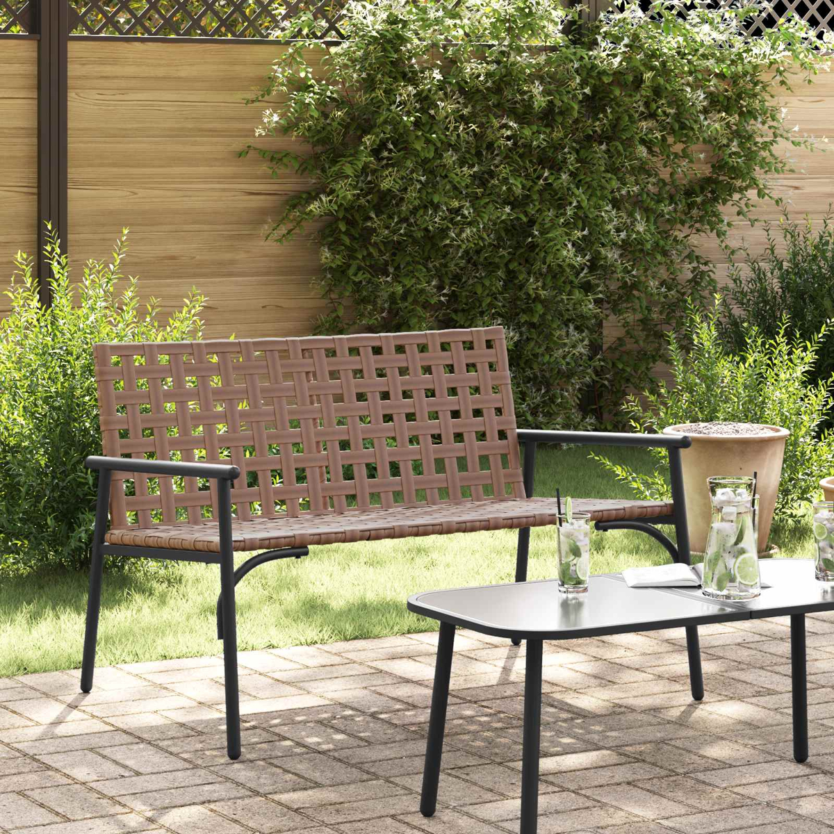 Eine braune, gewebte Bank steht auf einer Backstein-Terrasse mit einem kleinen Tisch und Getränken. Im Hintergrund befinden sich ein Holzzaun und Grün.