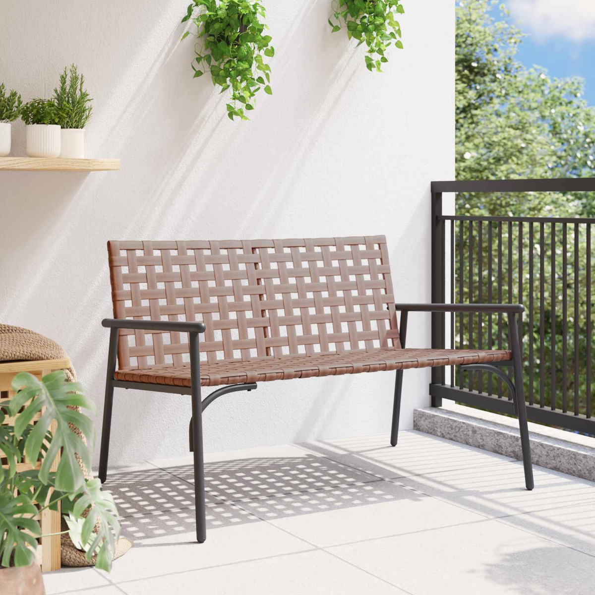 Eine braune, gewebte Bank mit schwarzem Metallrahmen auf einer weißen Terrasse. Grüne Pflanzen hängen darüber. Ein schwarzes Geländer ist im Hintergrund.