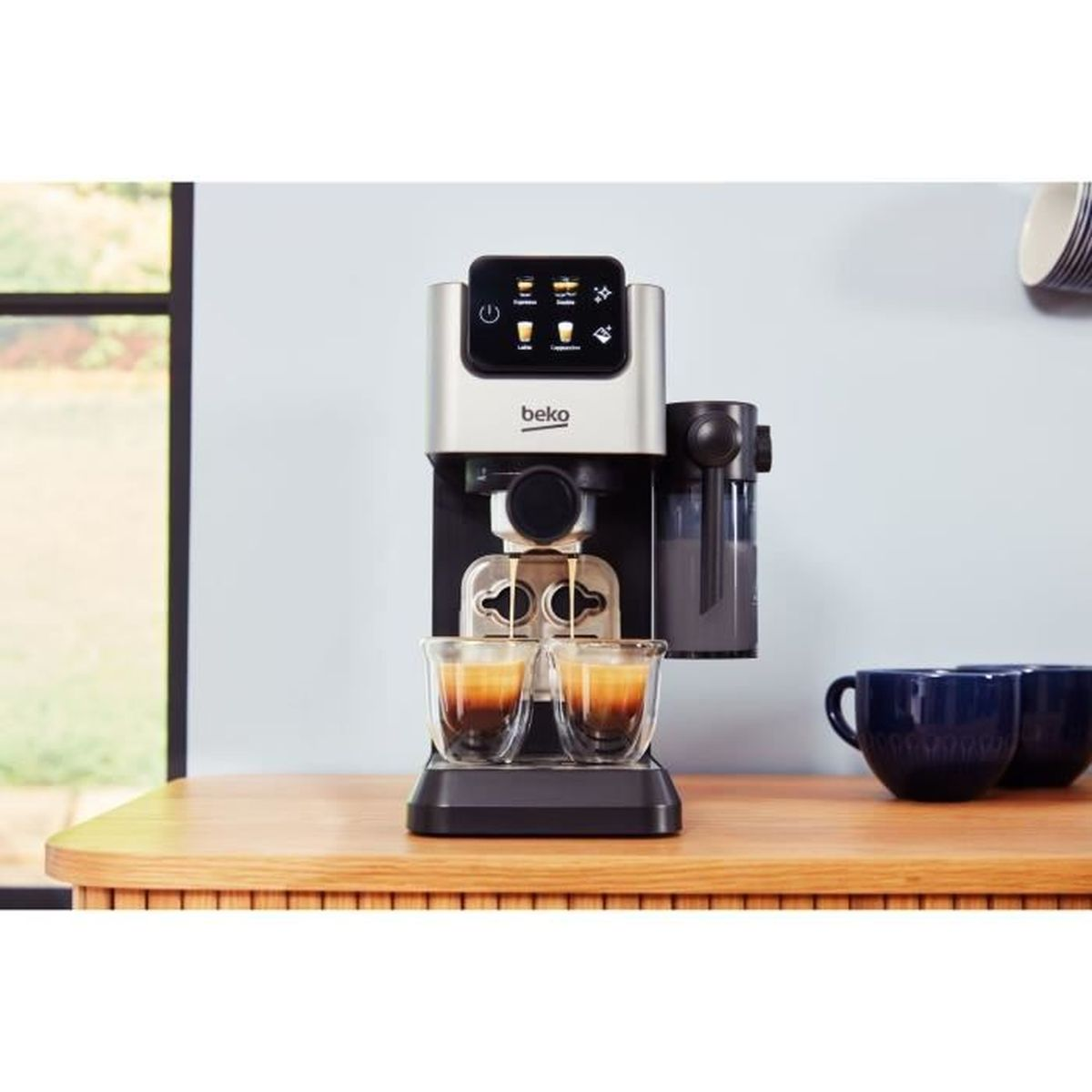 Espressomaschine, die Kaffee in zwei Gläser ausgibt. Maschine silber, Gläser klar, Tisch Holz, Tassen blau im Hintergrund.