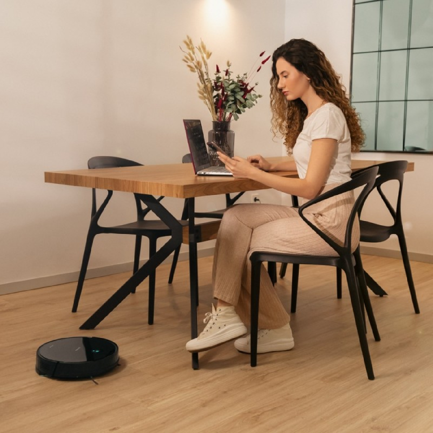 Una mujer sentada a una mesa usa un portátil y un teléfono. Un robot aspirador está en el suelo. Hay sillas, un jarrón y un espejo.