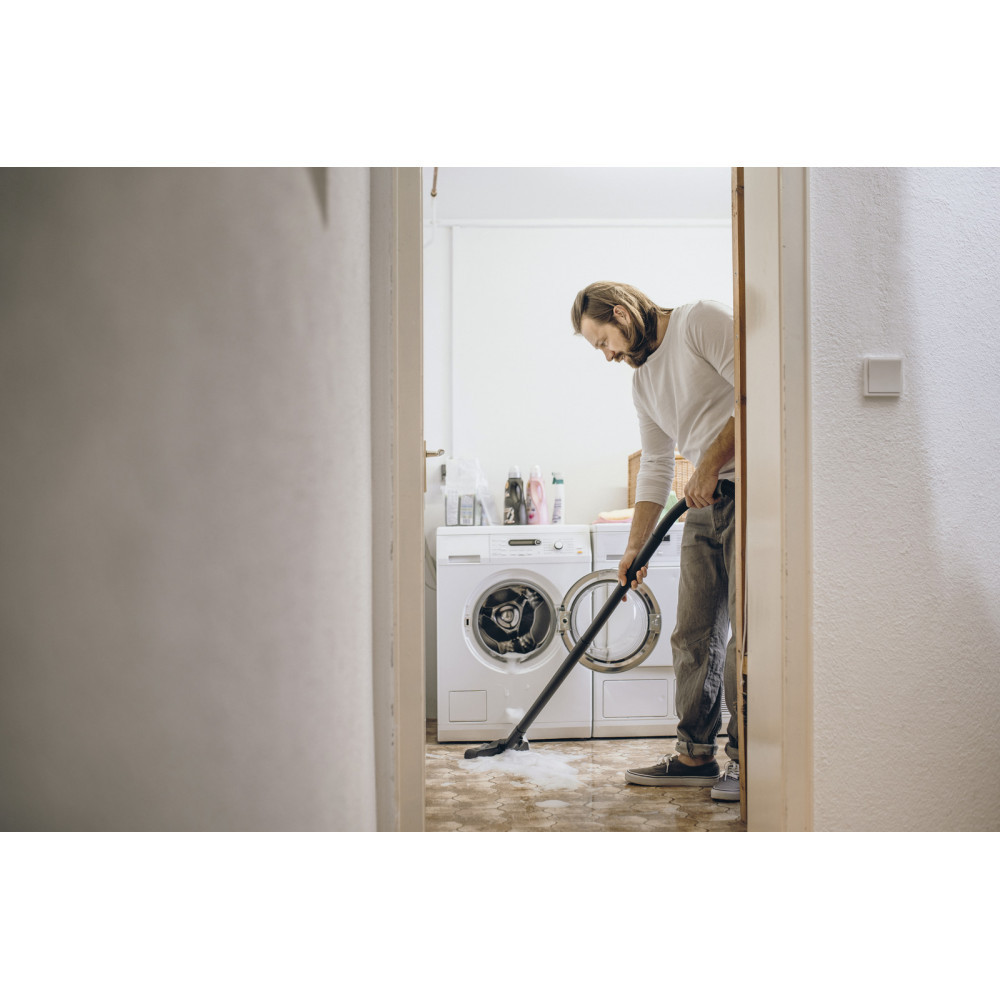 Mężczyzna odkurza pralnię. Ma na sobie białą koszulę i dżinsy. W tle dwie pralki.