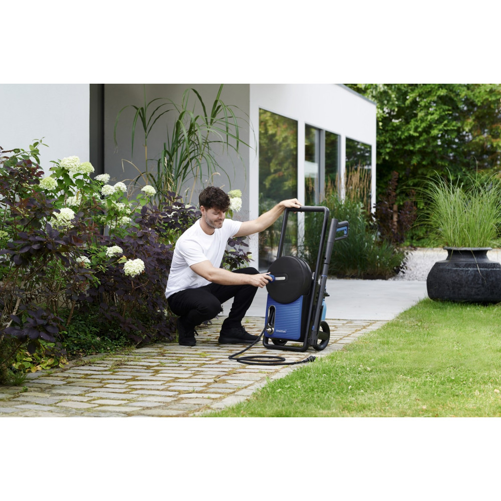 Mężczyzna w białej koszulce i czarnych spodniach obsługuje niebieską myjkę ciśnieniową na zewnątrz. Kuca.