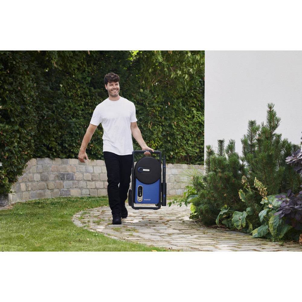 Mężczyzna niesie niebieską myjkę ciśnieniową po utwardzonej ścieżce. Zielone rośliny i kamienny mur w tle. Mężczyzna się uśmiecha.