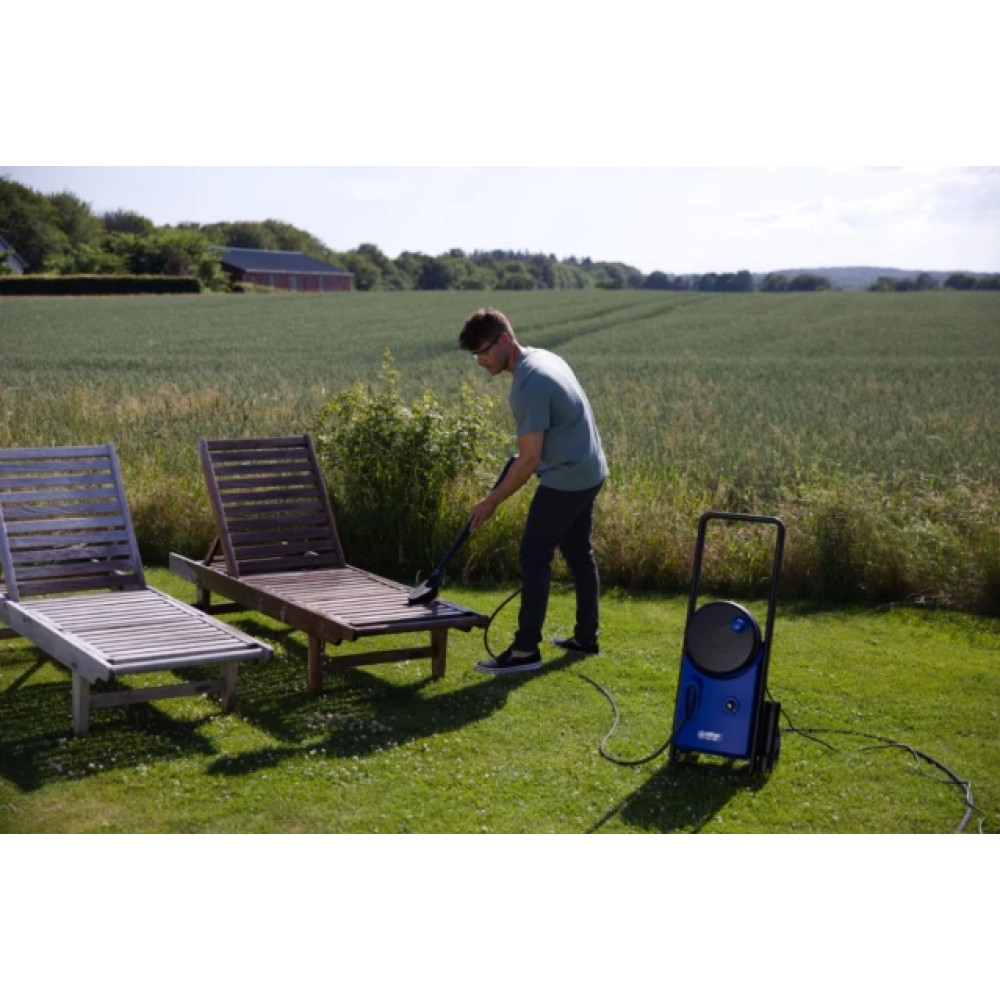 Mężczyzna używa myjki ciśnieniowej do drewnianego leżaka. Obok stoi kolejne krzesło. Zielone pole i drzewa w tle.