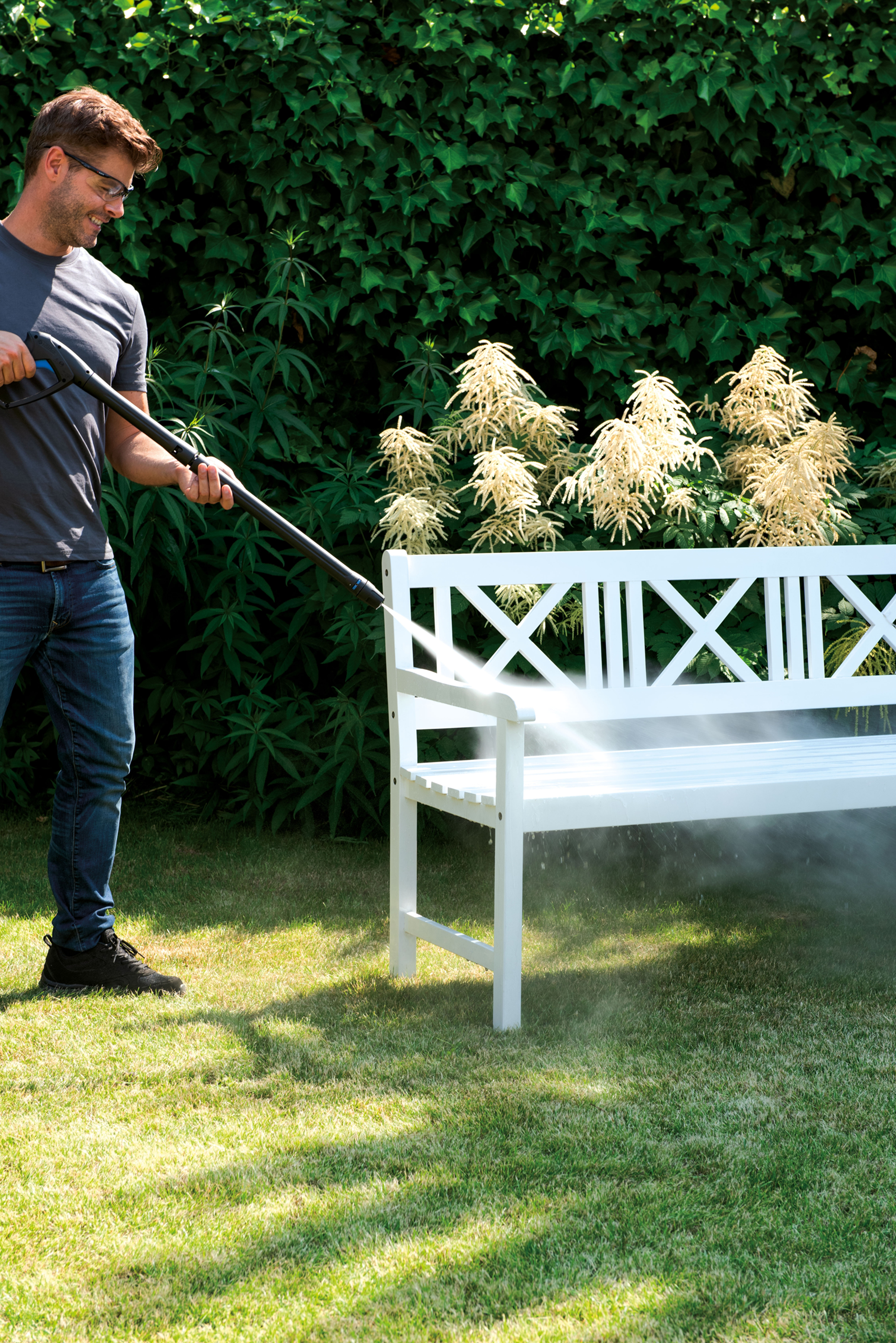 Mężczyzna używa myjki ciśnieniowej na białej ławce w ogrodzie. Zielone liście w tle.