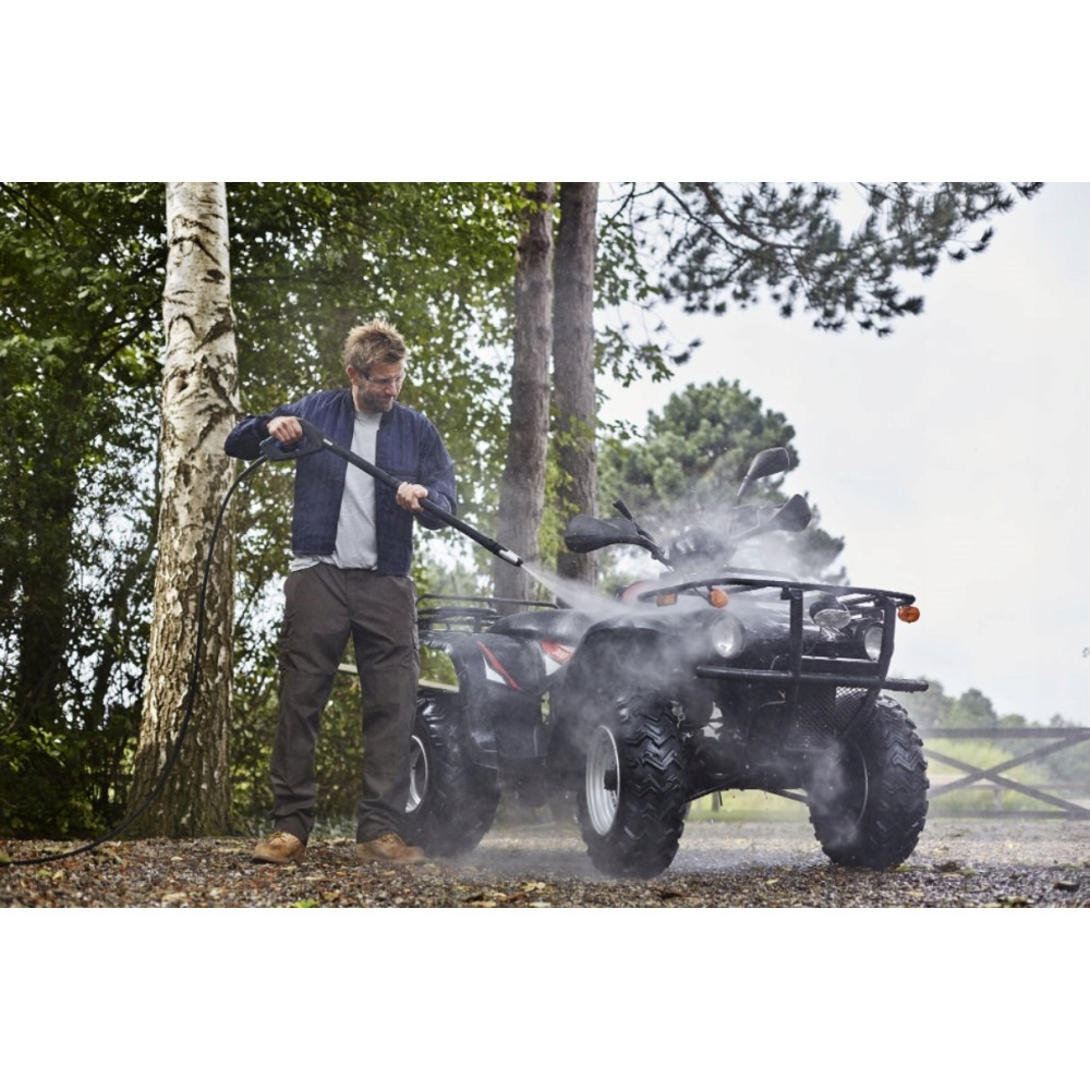Mężczyzna myje czarnego quada myjką ciśnieniową w zalesionym terenie. Woda spryskuje quada, tworząc mgłę.