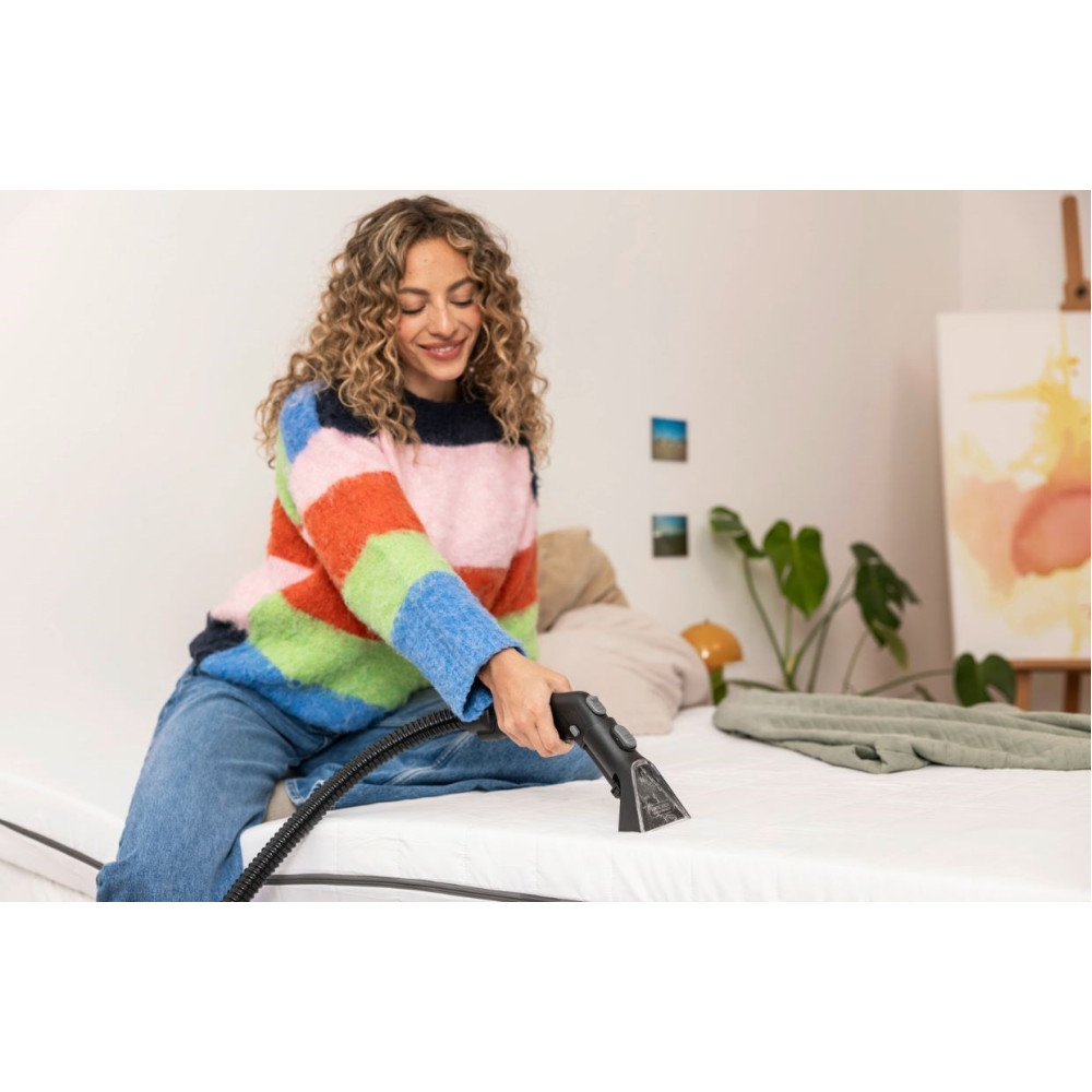 Kobieta w kolorowym swetrze czyści materac za pomocą narzędzia do czyszczenia. Scena sypialni ze sztuką, roślinami i pościelą.