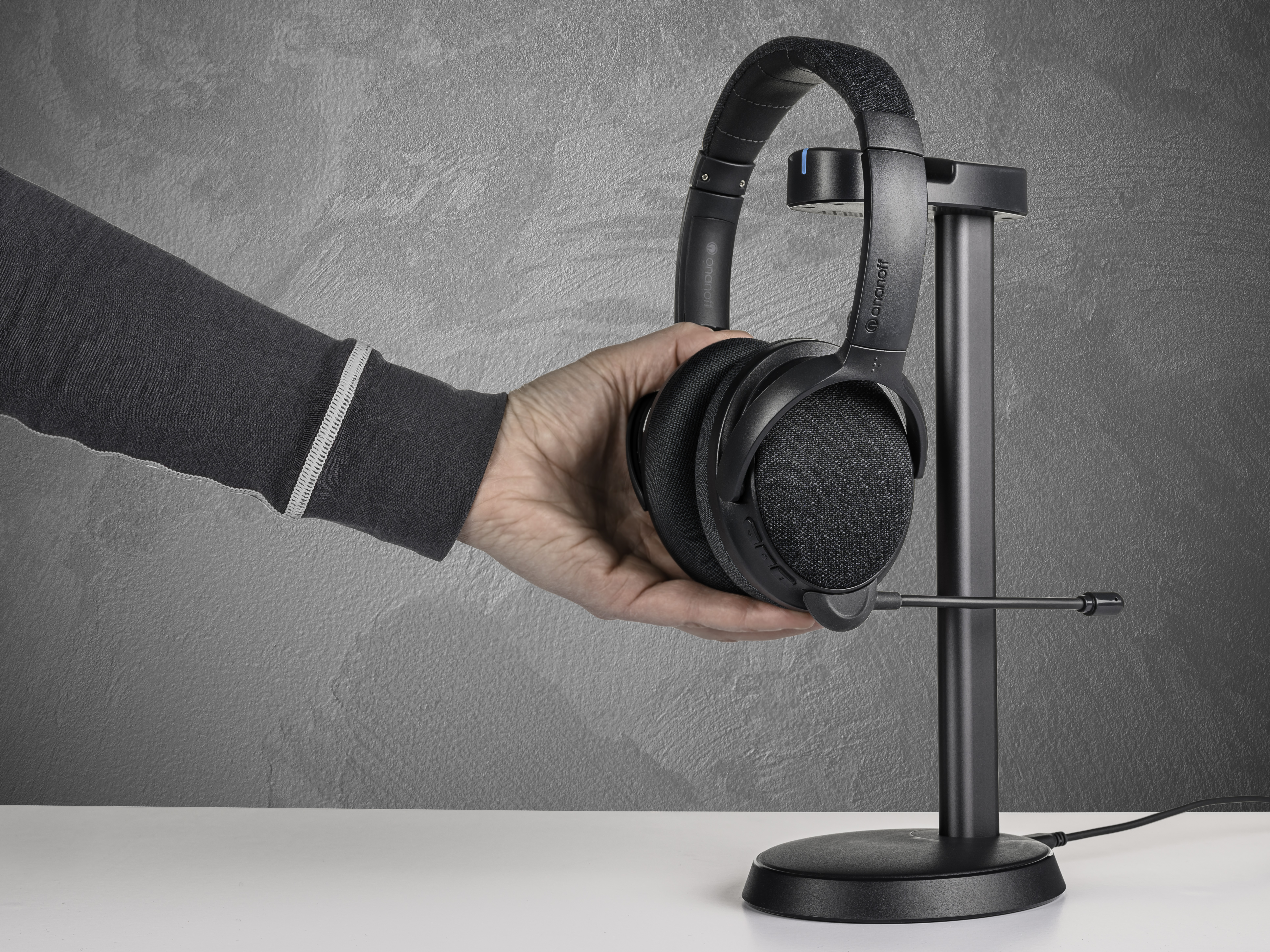 Eine Hand, die ein schwarzes Headset in der Nähe eines Ständers hält. Graue Wand im Hintergrund und ein weißer Tisch darunter.