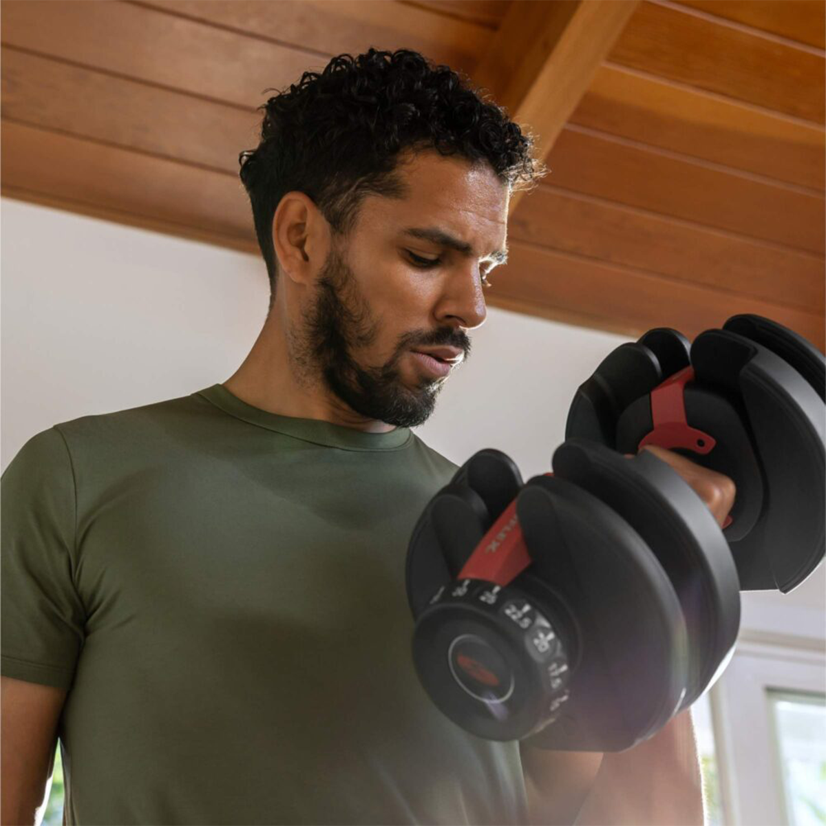 A man in a green shirt curls a black and red dumbbell. He is looking down. The background shows wood paneling.