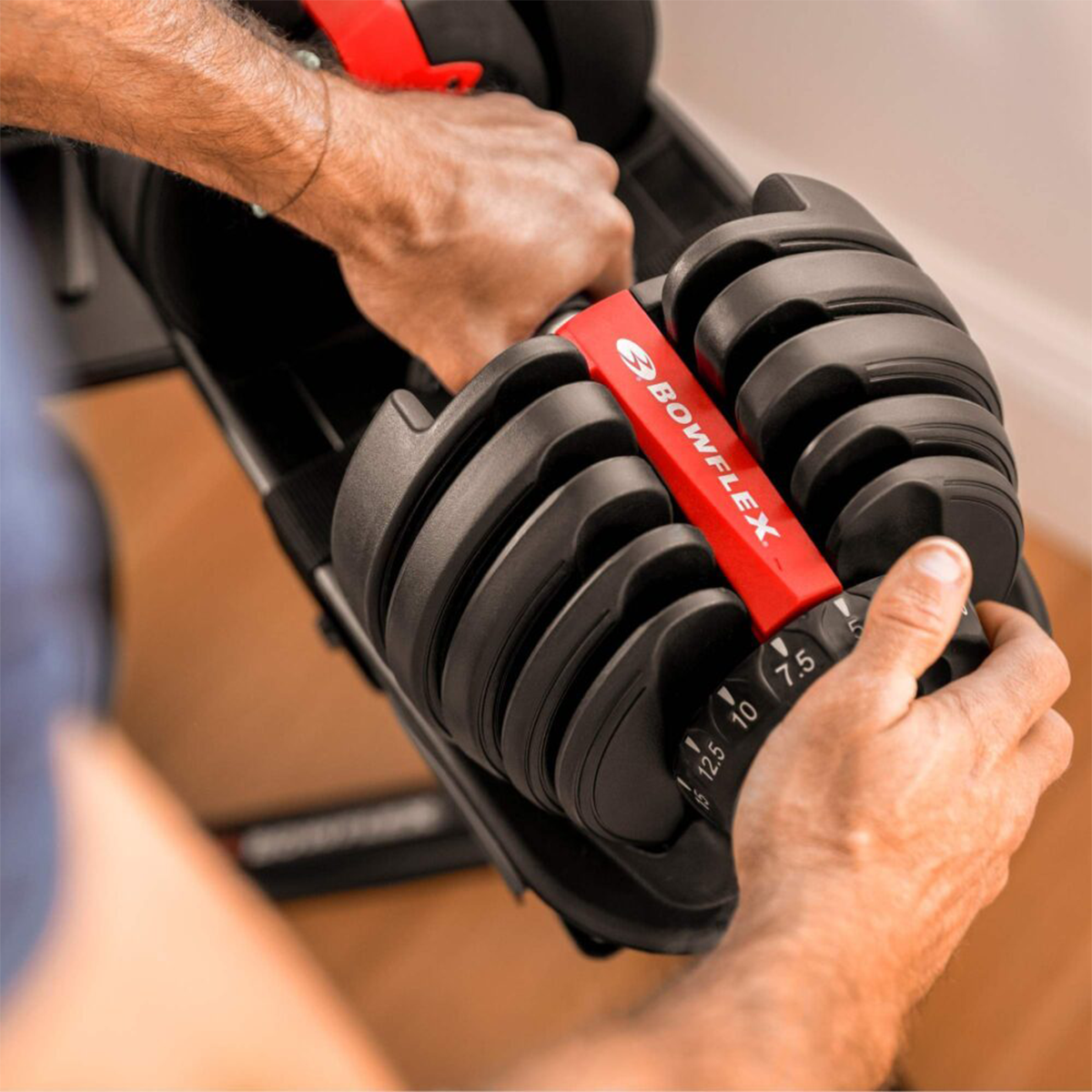 A person adjusting a Bowflex dumbbell. Black weights with red accents are visible. The person's hands are on the weight selector.