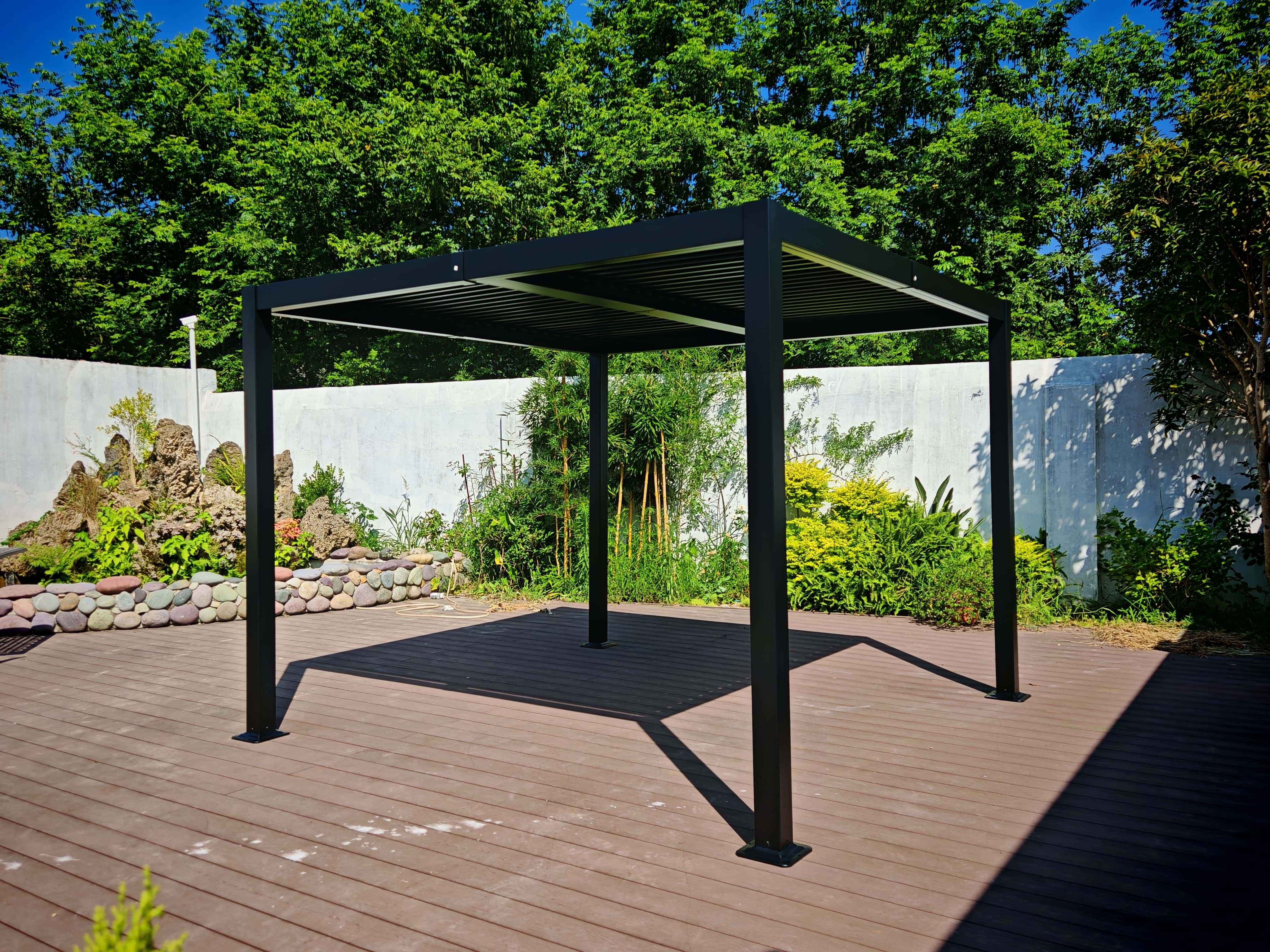 Schwarze Pergola auf Holzterrasse. Grüne Bäume im Hintergrund. Ein Garten mit grünen Pflanzen befindet sich an der Seite.
