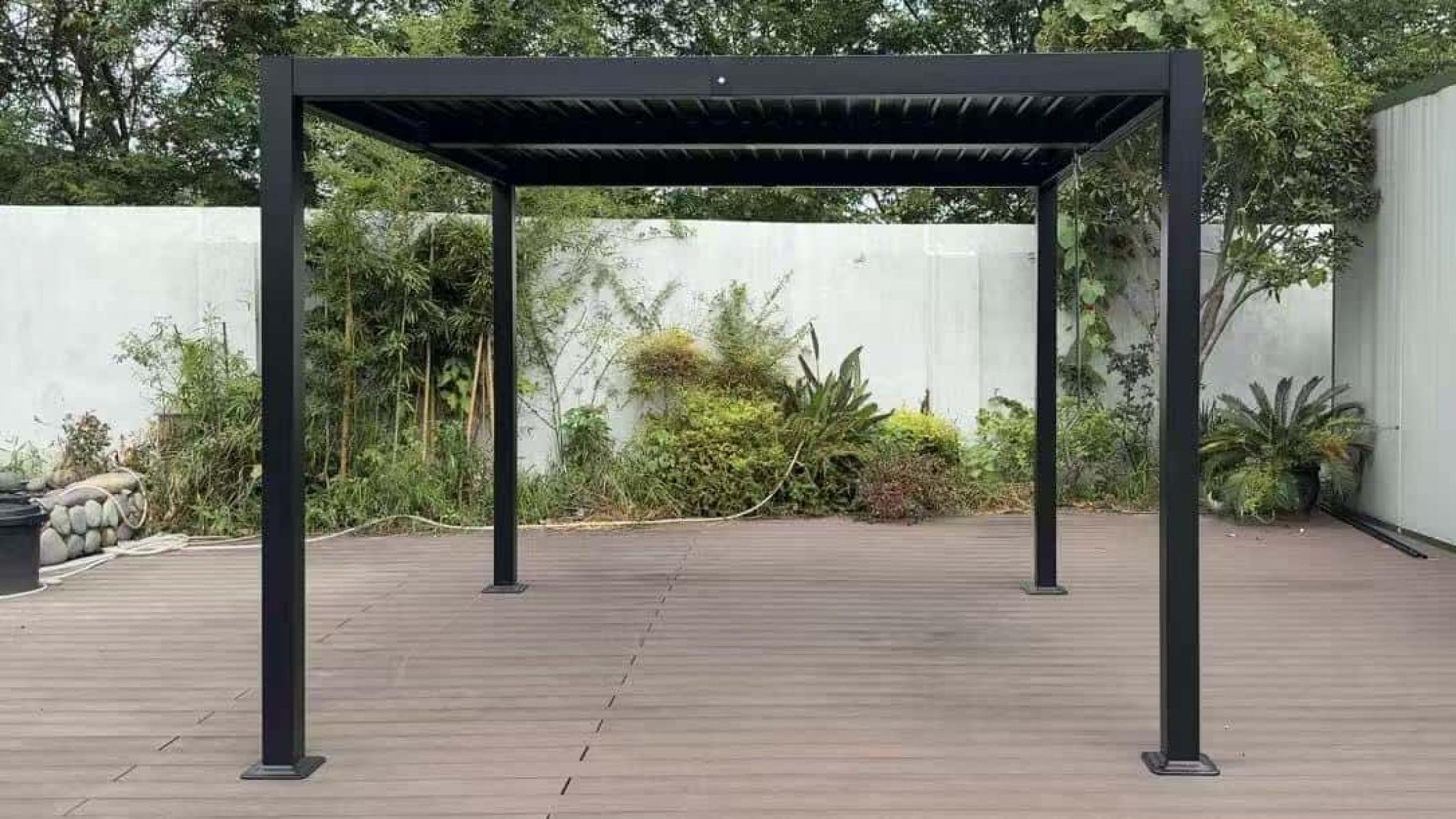 Eine schwarze Pergola steht auf einem Holzdeck. Grüne Pflanzen befinden sich im Hintergrund vor einer weißen Wand.